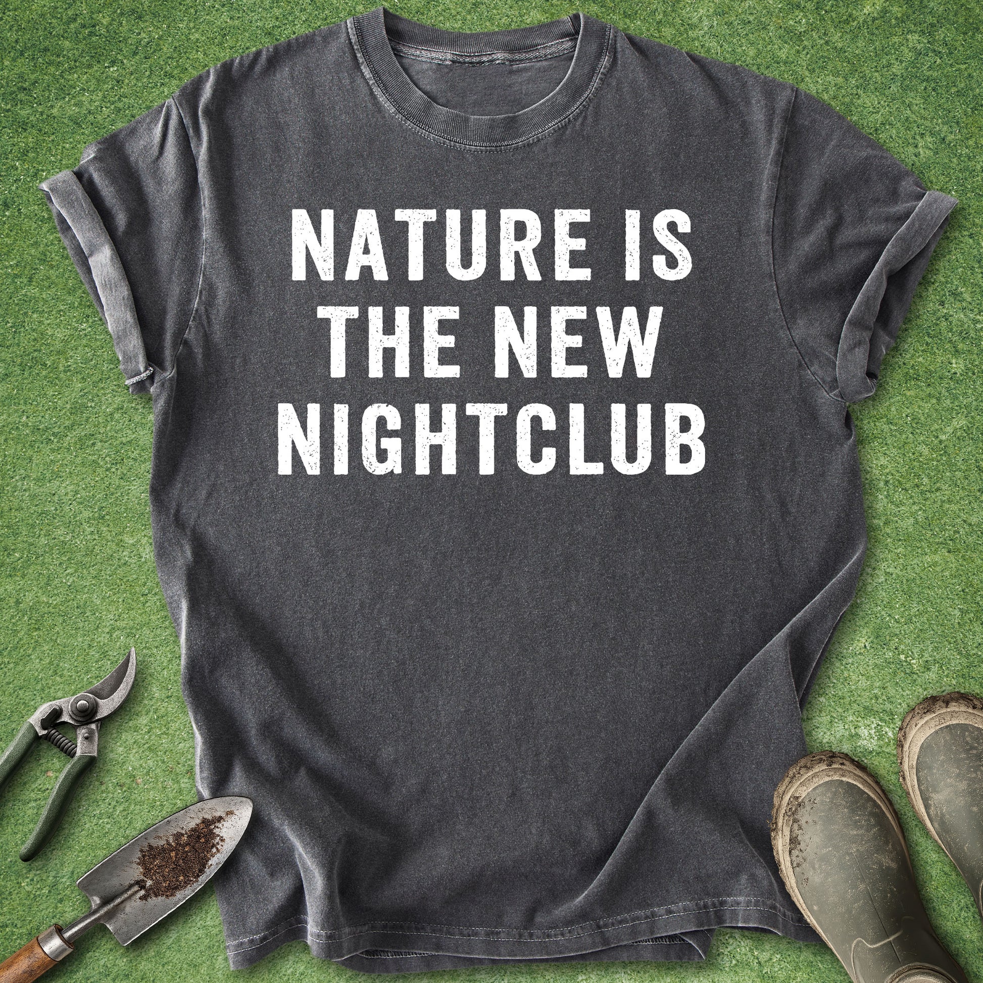 A dark charcoal gray graphic t-shirt with the text 'NATURE IS THE NEW NIGHTCLUB' printed in bold white capital letters, laid flat on green grass with gardening tools and boots nearby.