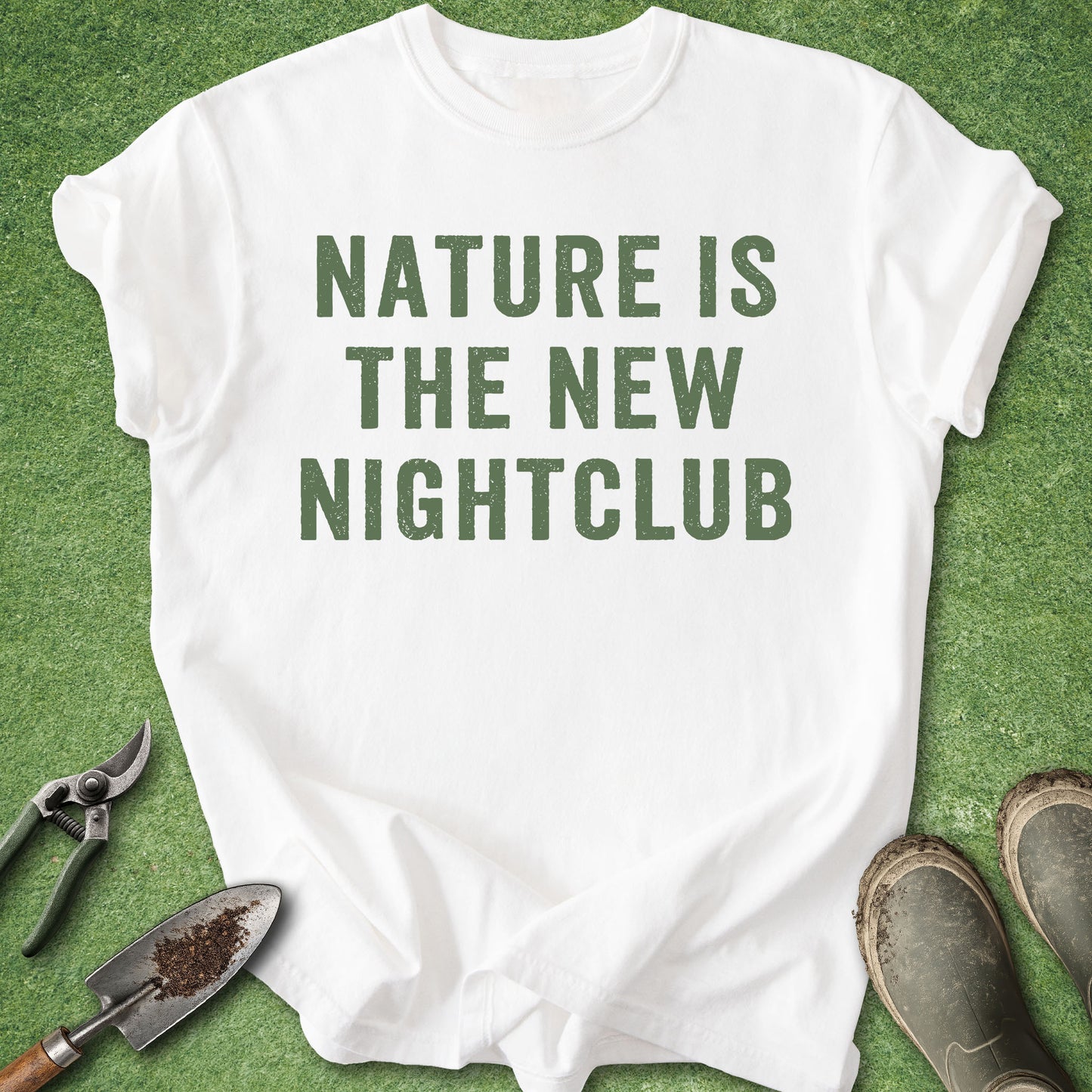 A white short-sleeve T-shirt laid flat on green grass-like ground, with large dark green text reading “NATURE IS THE NEW NIGHTCLUB.” Gardening tools and muddy boots are placed around it as styling props.