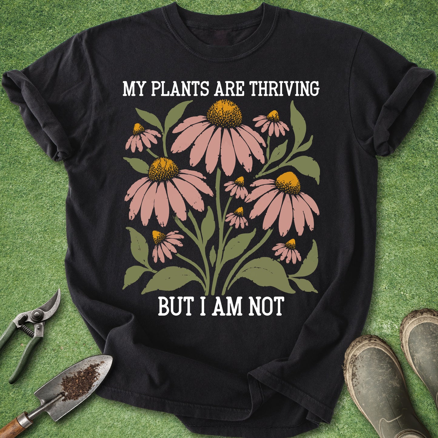 A black short-sleeve graphic T-shirt laid flat on green artificial turf, with white text reading 'MY PLANTS ARE THRIVING BUT I AM NOT' and a floral print of pink coneflowers with green leaves. Gardening tools and muddy boots are placed around the shirt as styled props.