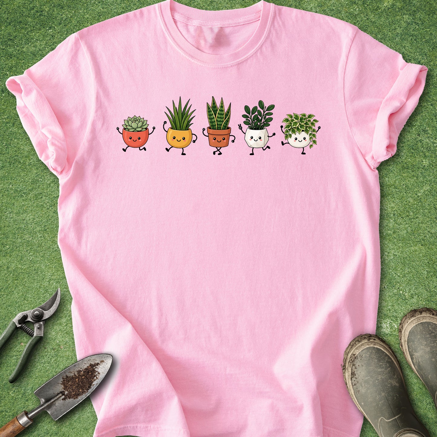 A pink short-sleeve t-shirt laid flat on green grass, featuring a graphic of five cute, smiling cartoon potted plants in different colors. Gardening props, including pruning shears, a trowel, and gardening boots, are arranged around the shirt.