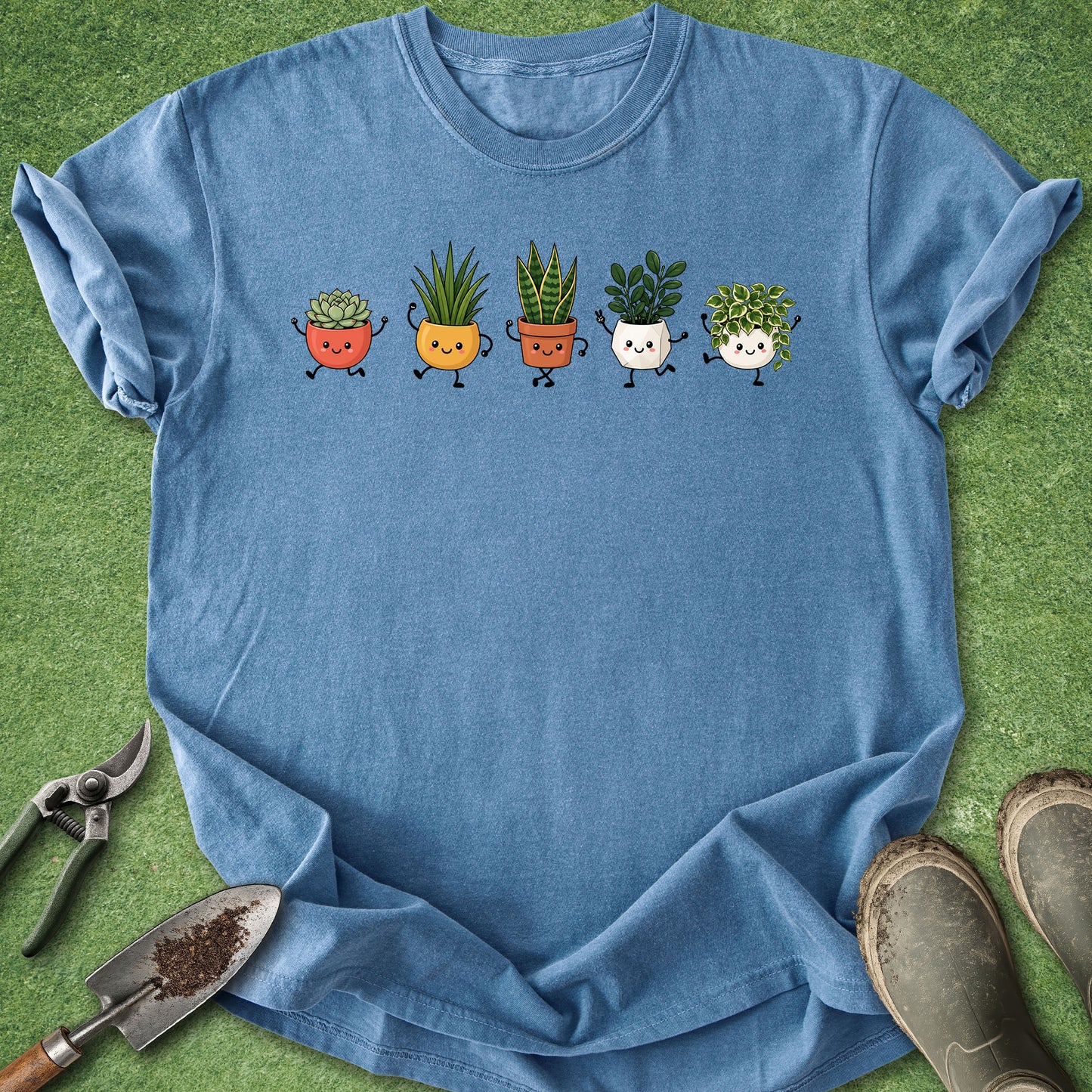 A blue short-sleeve crewneck t-shirt laid flat on a green grass-like surface, featuring a graphic of five cute cartoon potted plants across the chest. Gardening props including pruning shears, a small trowel, and muddy boots are visible around the shirt.