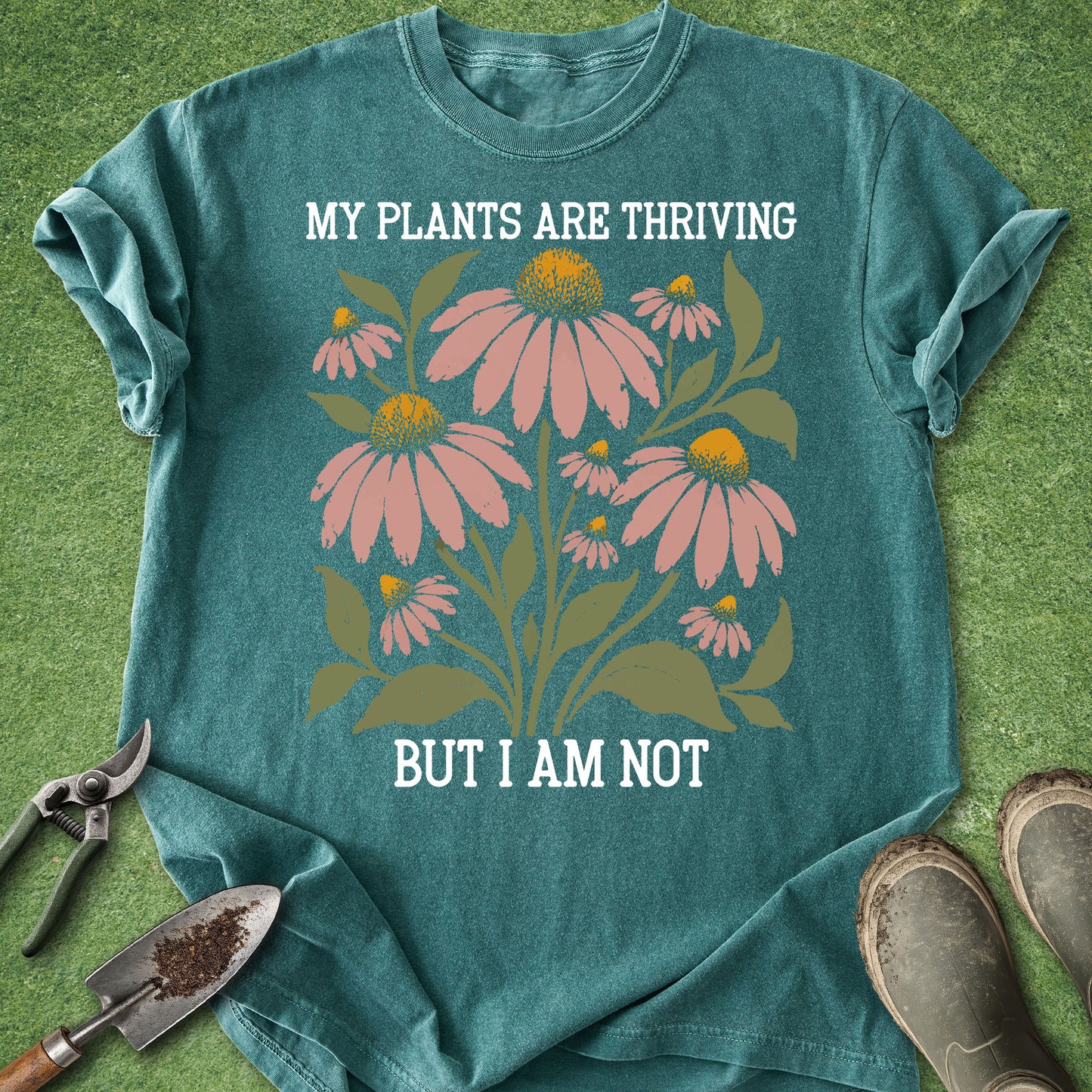 A teal graphic t-shirt laid flat on a green grass-like surface, with gardening tools and boots around it. The shirt has a floral print of pink coneflowers with yellow centers and white text reading 'MY PLANTS ARE THRIVING BUT I AM NOT'.