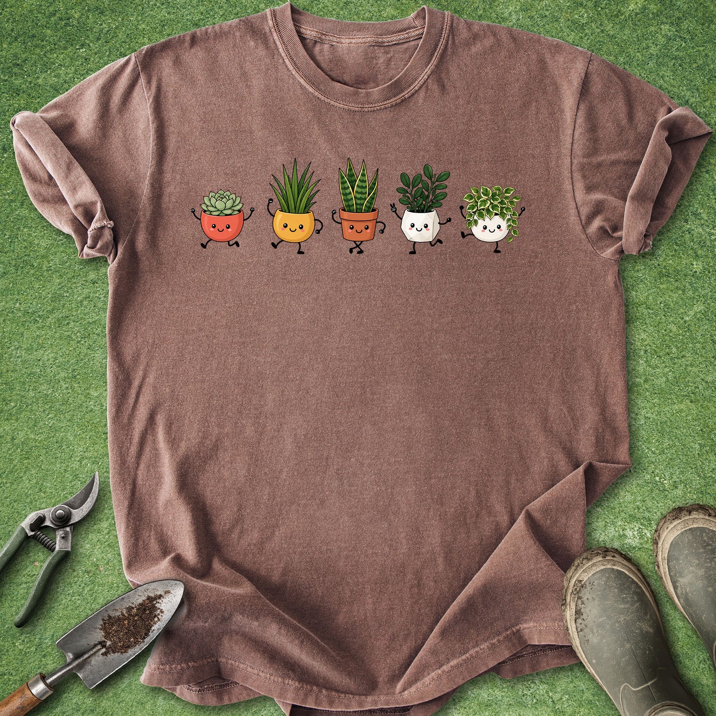 A brown short-sleeve t-shirt laid flat on green artificial turf, featuring a graphic of five cute cartoon potted plants in different colors. Gardening props, including pruning shears, a trowel with soil, and muddy boots, are arranged around the shirt.
