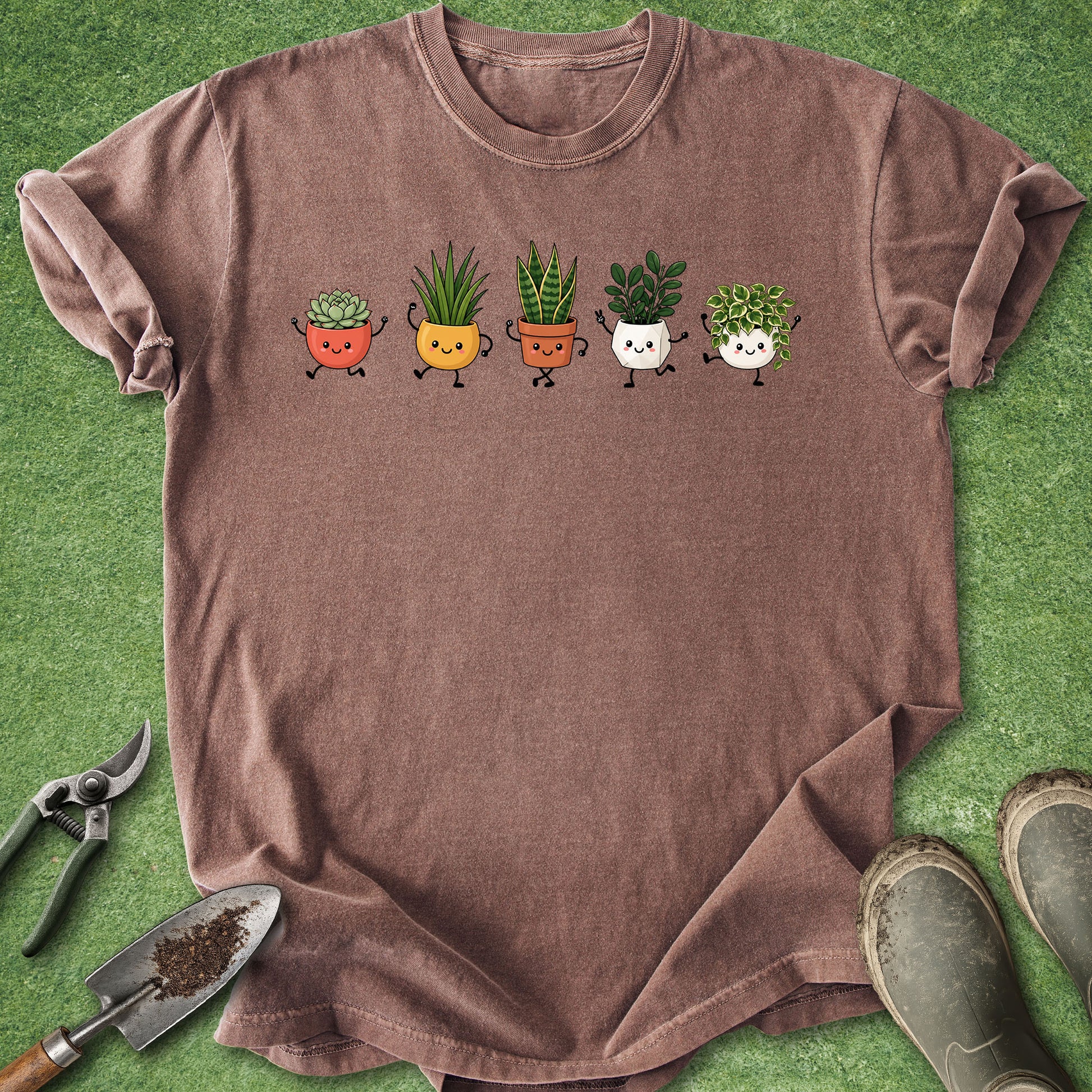 A brown short-sleeve t-shirt laid flat on green artificial turf, featuring a graphic of five cute cartoon potted plants in different colors. Gardening props, including pruning shears, a trowel with soil, and muddy boots, are arranged around the shirt.