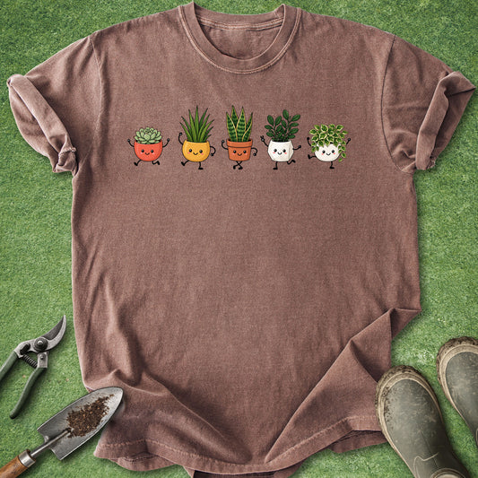 A brown short-sleeve t-shirt laid flat on green artificial turf, featuring a graphic of five cute cartoon potted plants in different colors. Gardening props, including pruning shears, a trowel with soil, and muddy boots, are arranged around the shirt.