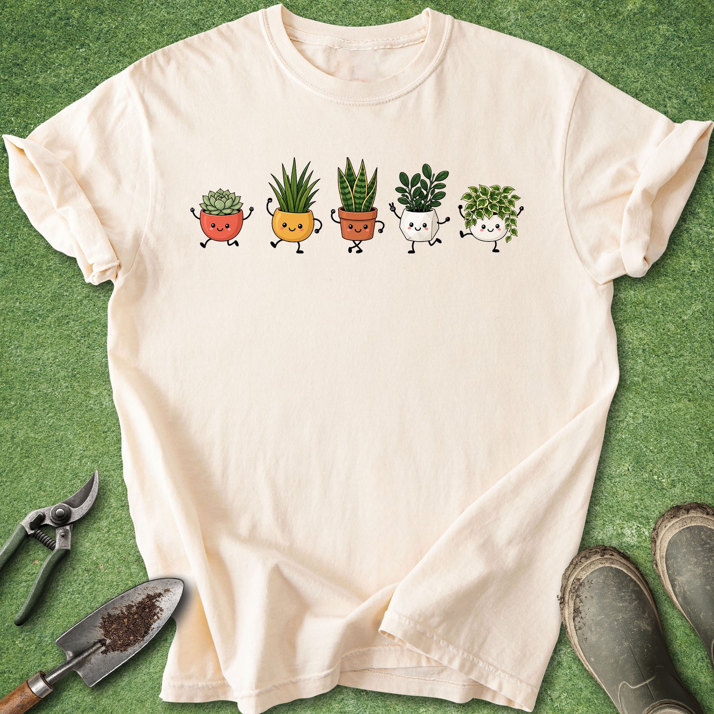 A cream-colored short-sleeve t-shirt laid flat on a green grass-like surface, featuring a graphic of five cute cartoon potted plants in different colors. Gardening tools and muddy boots are placed around the shirt as props.