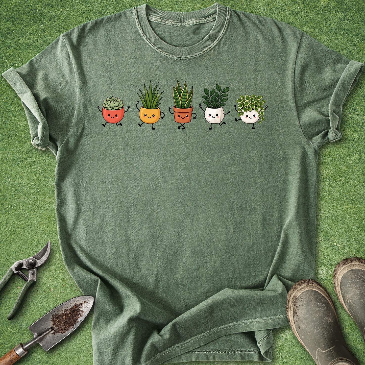 A heathered sage green t-shirt laid flat on a green grass-like surface, featuring a graphic of five cute cartoon potted plants in different colors. Gardening props, including pruning shears, a trowel, and boots, are arranged around the shirt.