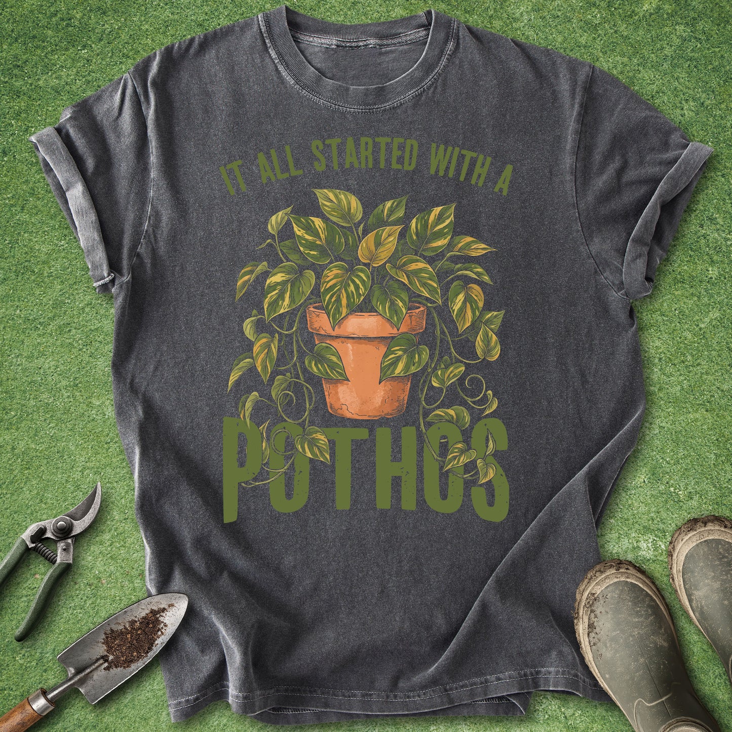 A dark charcoal gray t-shirt with a green and terracotta graphic of a pothos plant and the text 'IT ALL STARTED WITH A POTHOS', styled in a flat-lay on artificial green grass with gardening tools and boots.