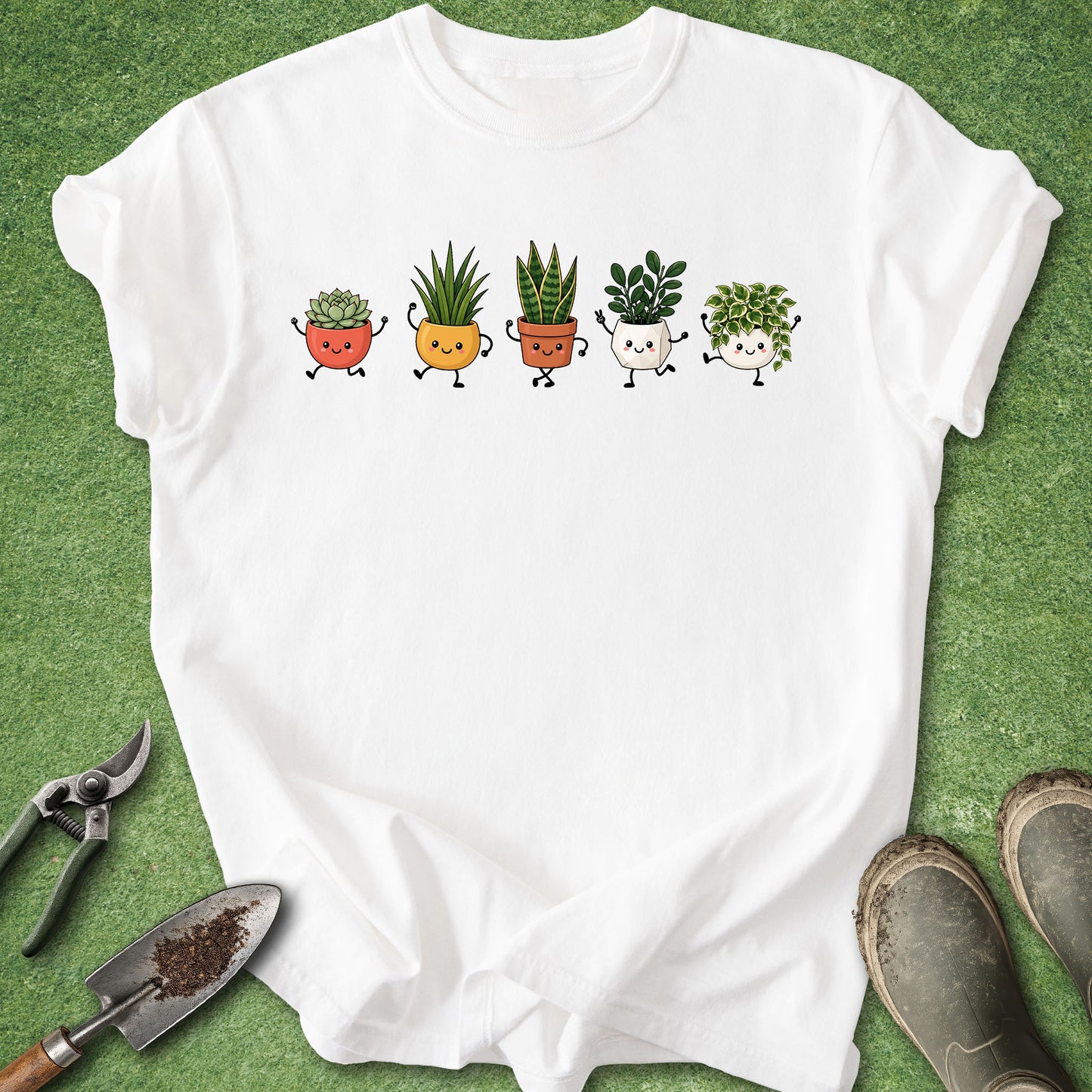 A white short-sleeve T-shirt laid flat on green grass-like artificial turf, featuring a row of five cute cartoon potted plants across the chest. Gardening tools and muddy boots are placed around the shirt as styling props.