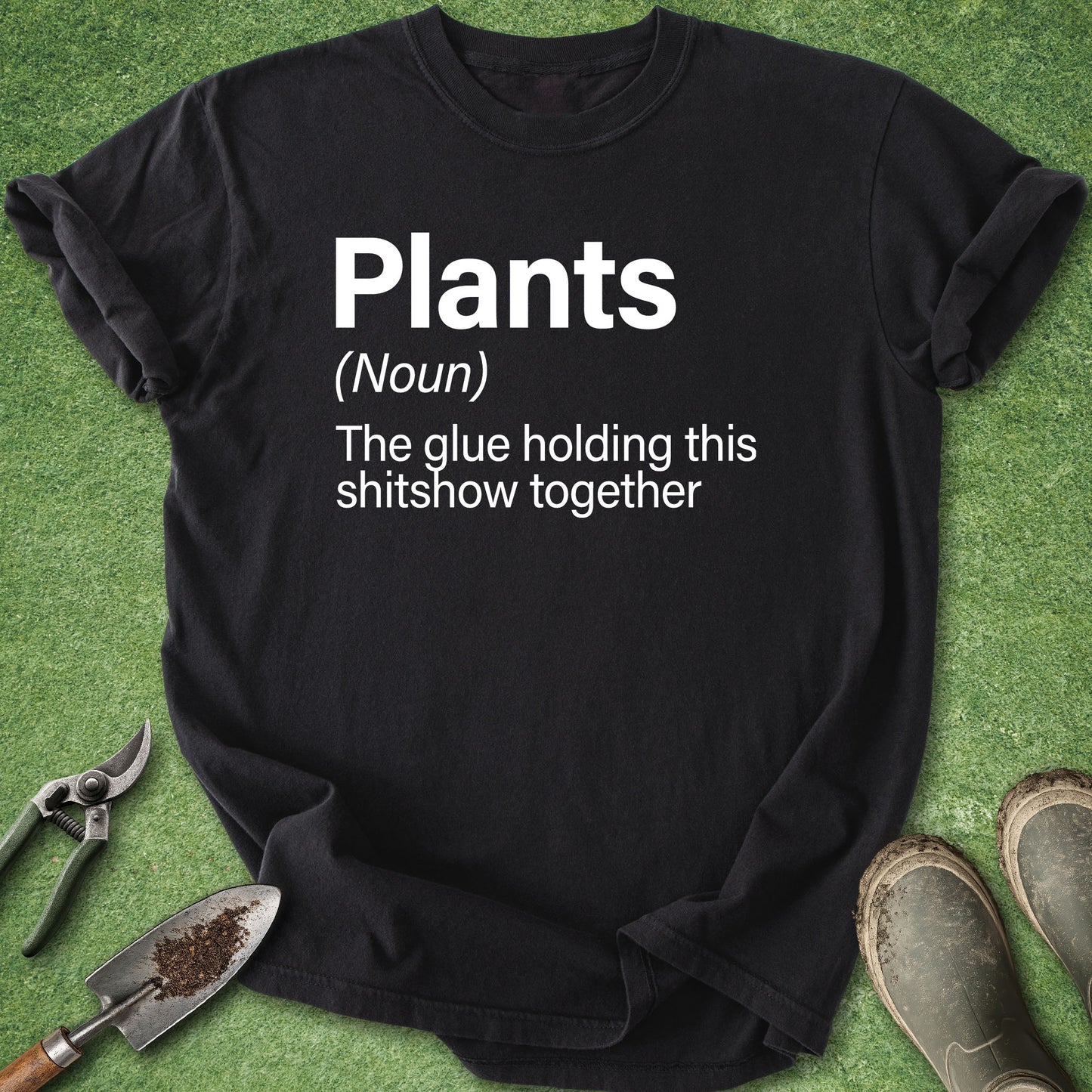 A black graphic t-shirt laid flat on green grass-like artificial turf, with white printed text reading 'Plants (Noun) The glue holding this shitshow together.' Gardening props including pruning shears, a trowel, and boots are placed around the shirt.