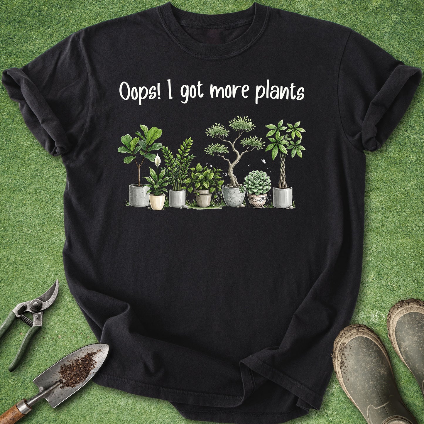 A black short-sleeve graphic T-shirt laid flat on green grass-like background, with white text reading 'Oops! I got more plants' above a row of illustrated potted houseplants. Gardening props including pruning shears, a trowel, and boots are partially visible around the shirt.