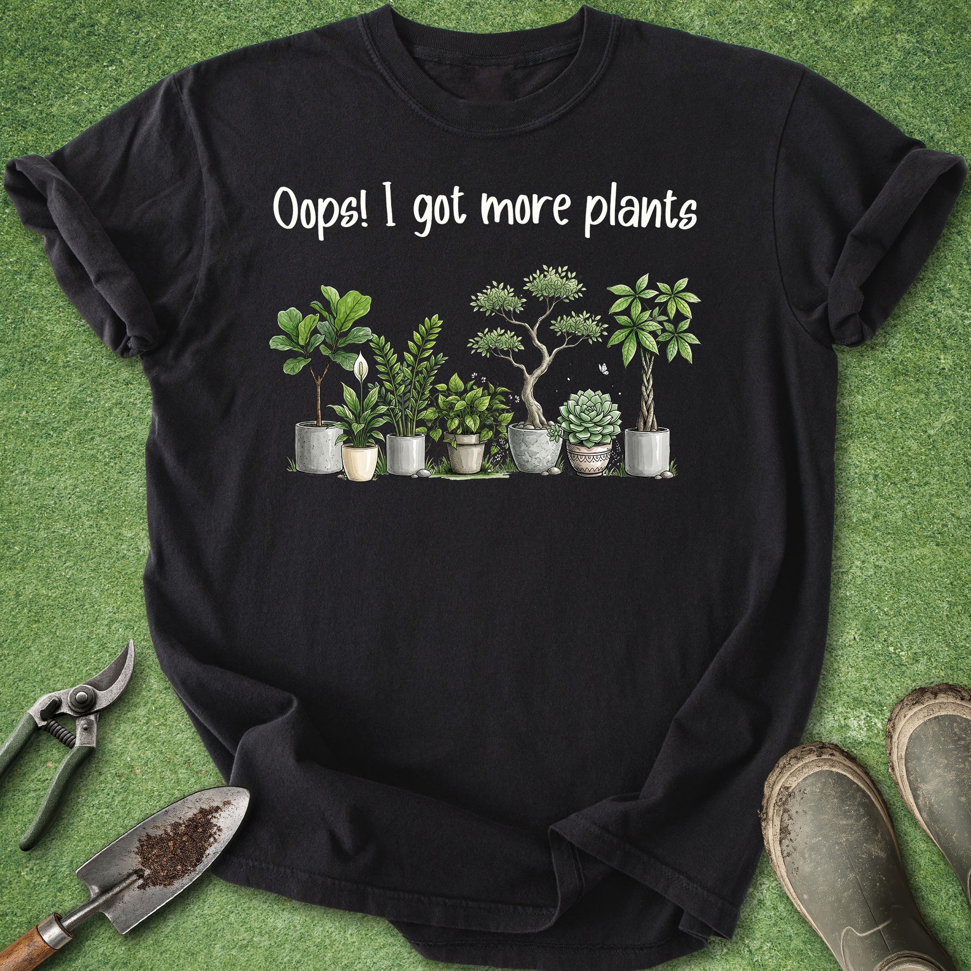 A black short-sleeve graphic T-shirt laid flat on green grass-like background, with white text reading 'Oops! I got more plants' above a row of illustrated potted houseplants. Gardening props including pruning shears, a trowel, and boots are partially visible around the shirt.