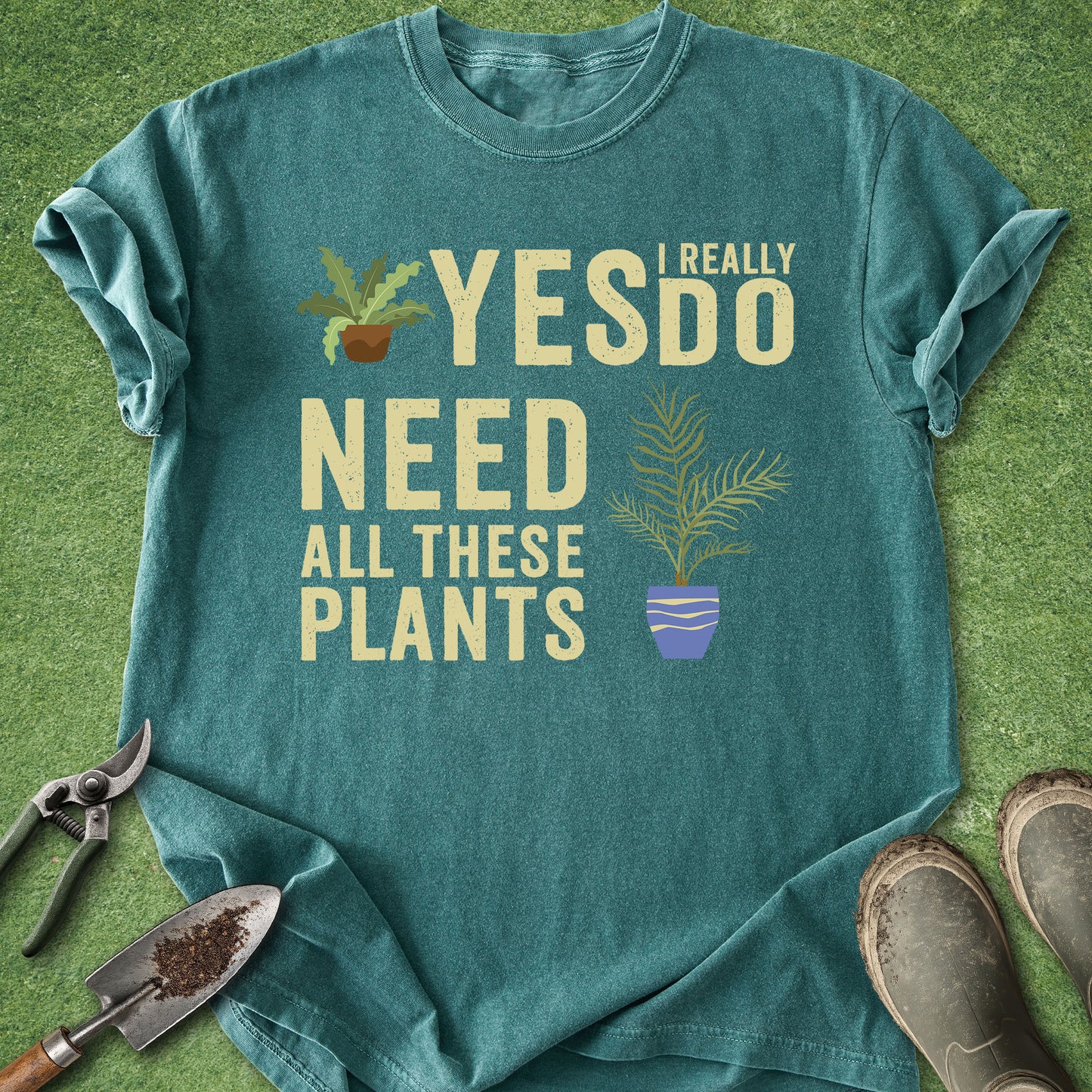 Teal t-shirt with 'I really do need all these plants' text and plant graphics on a grass background.