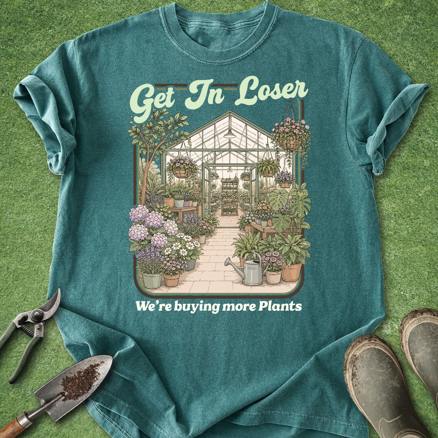 A teal graphic t-shirt laid flat on a green grass-like surface, featuring a greenhouse and potted plants illustration with the text 'Get In Loser' and 'We're buying more Plants'. Gardening props including pruning shears, a trowel, and boots are arranged around the shirt.