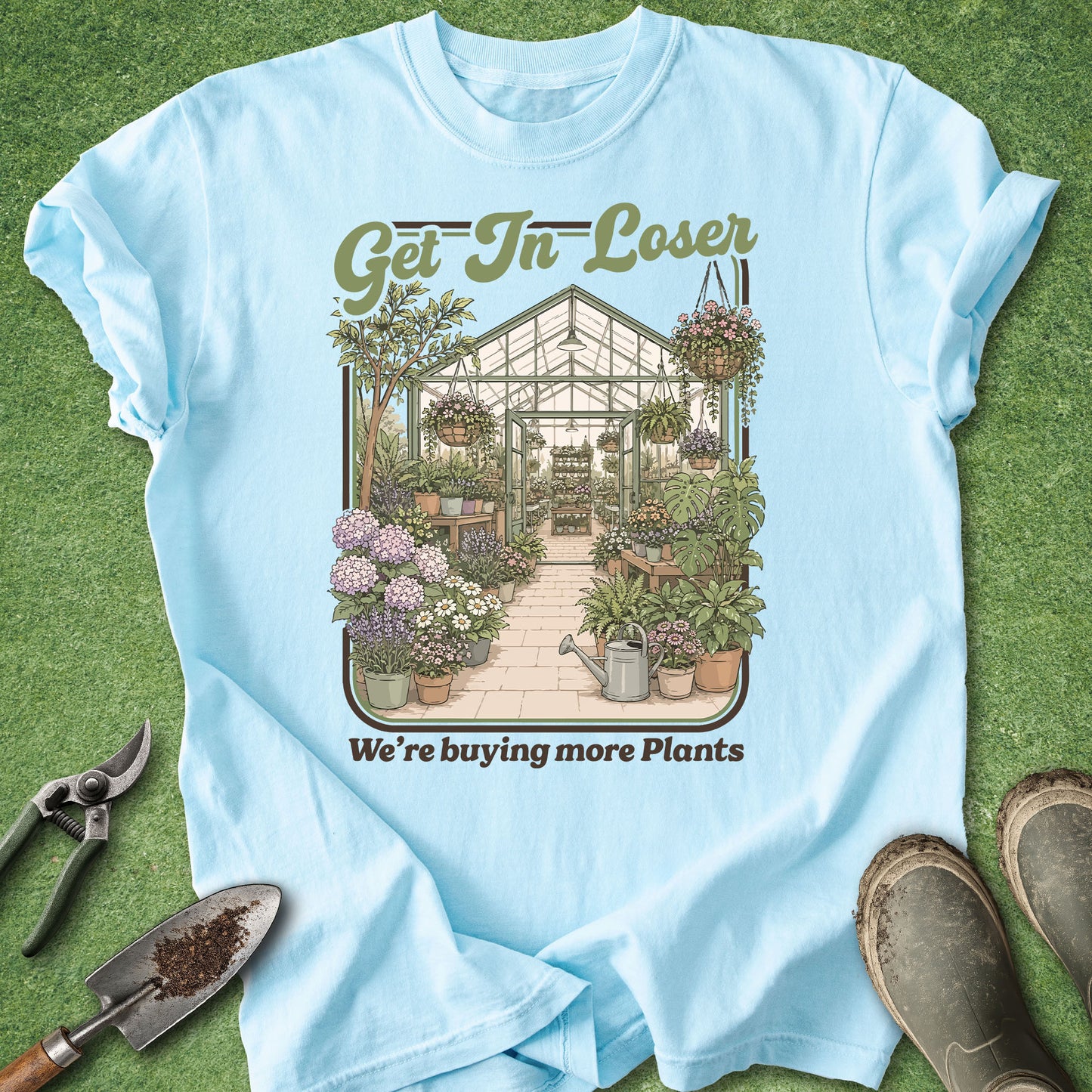 A light blue graphic t-shirt laid flat on green grass, surrounded by gardening props including pruning shears, a trowel, and work boots. The shirt features a central illustration of a lush greenhouse filled with plants, with the text 'Get In Loser' above and 'We're buying more Plants' below.