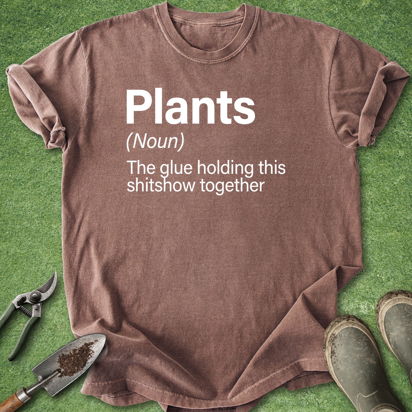 A brown short-sleeve graphic T-shirt laid flat on green grass-like ground, with white printed text reading 'Plants (Noun) The glue holding this shitshow together.' Gardening tools and a pair of boots are partially visible around the shirt as styling props.