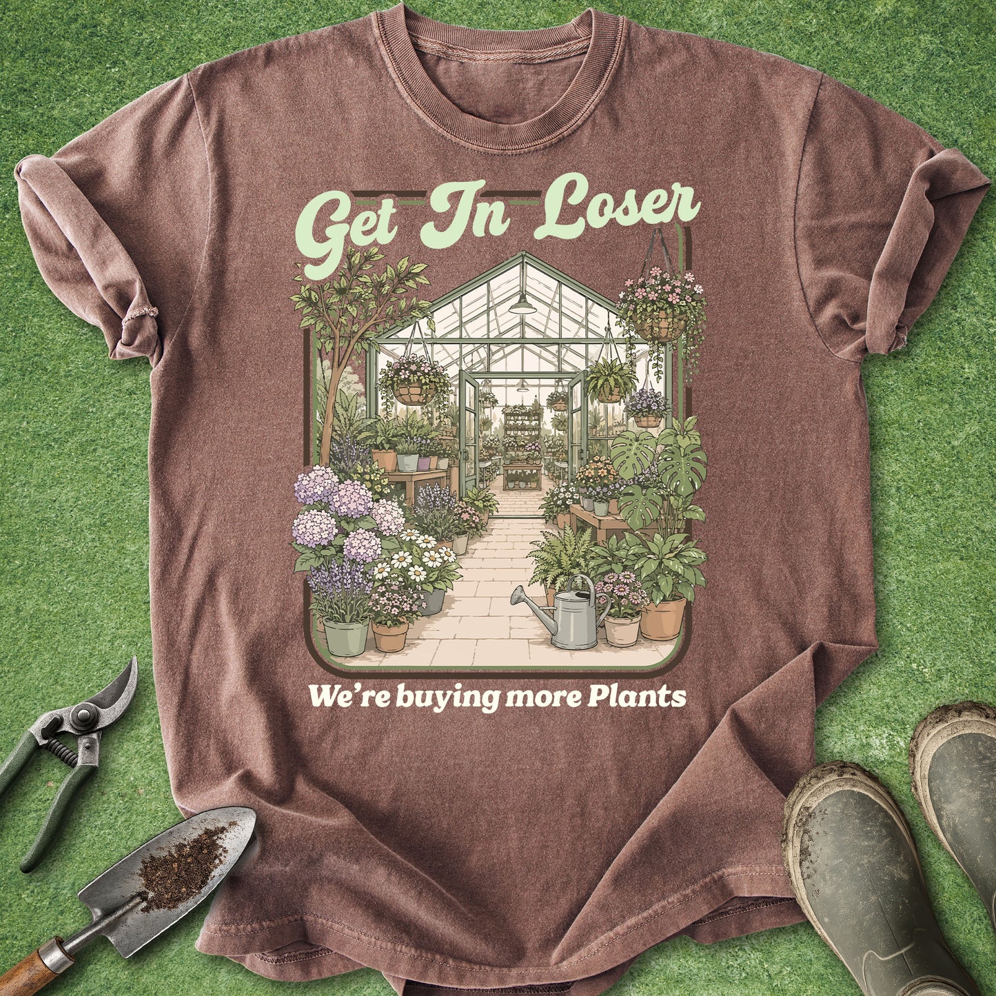 A brown graphic t-shirt laid flat on green grass-like background, with gardening tools and boots around it. The shirt has a greenhouse and plants illustration with the text 'Get In Loser' and 'We're buying more Plants'.