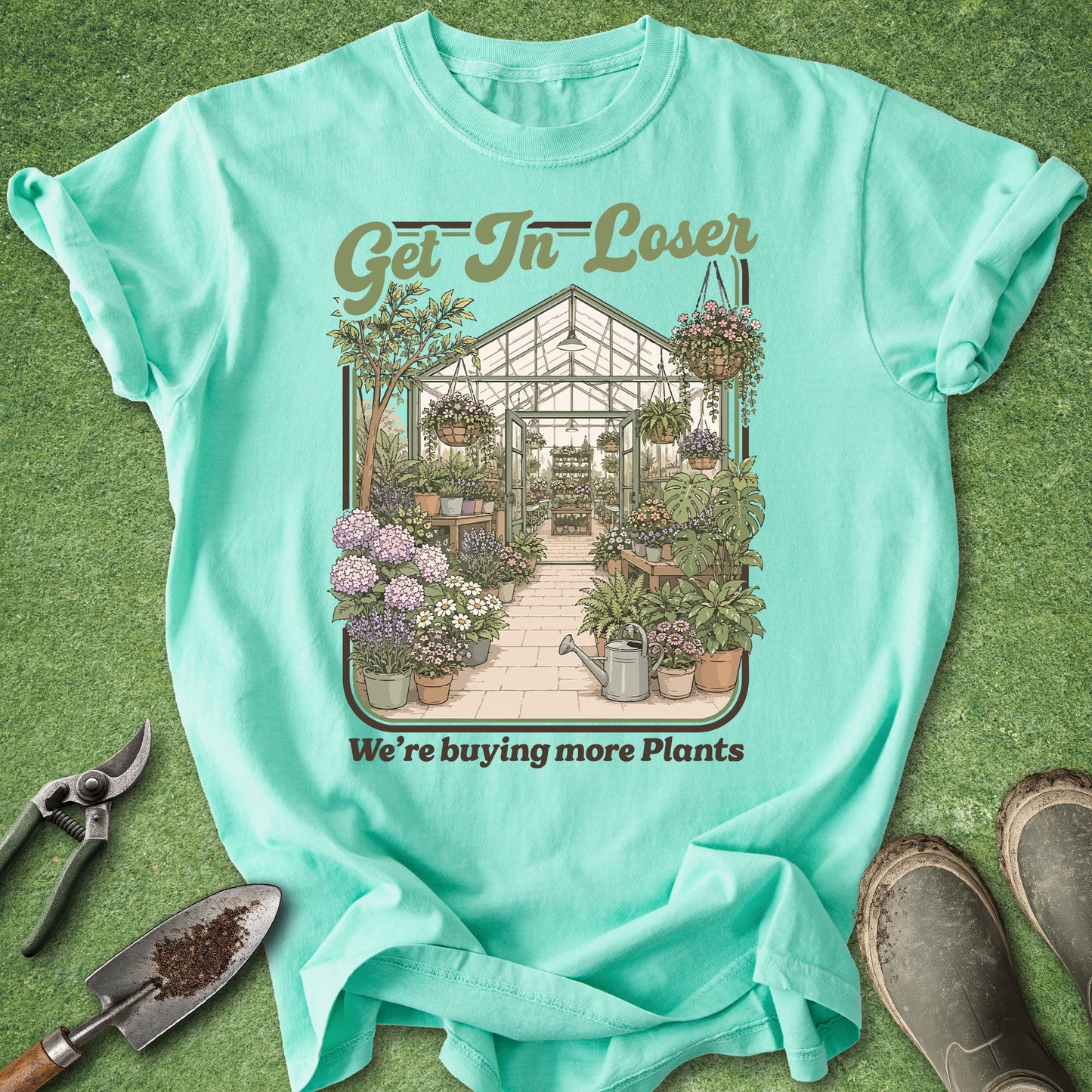 A mint green graphic t-shirt laid flat on a green grass-like surface, surrounded by gardening props including pruning shears, a trowel, and work boots. The shirt features a detailed illustration of a greenhouse filled with plants and the text 'Get In Loser' and 'We're buying more Plants'.