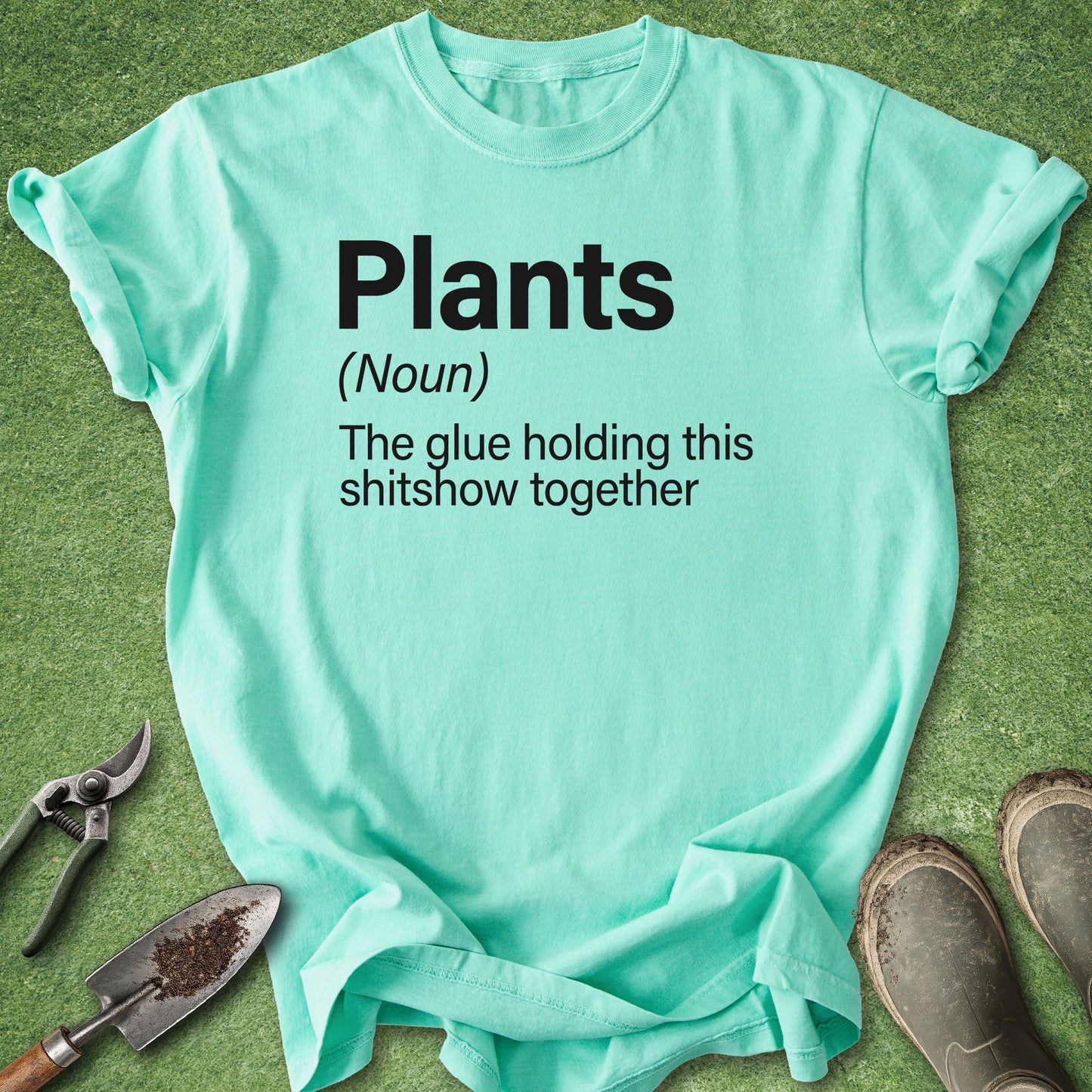 A mint green graphic t-shirt laid flat on artificial grass with gardening props, including pruning shears, a trowel, and muddy boots. The shirt features a black printed text definition of the word 'Plants' with a humorous definition underneath.