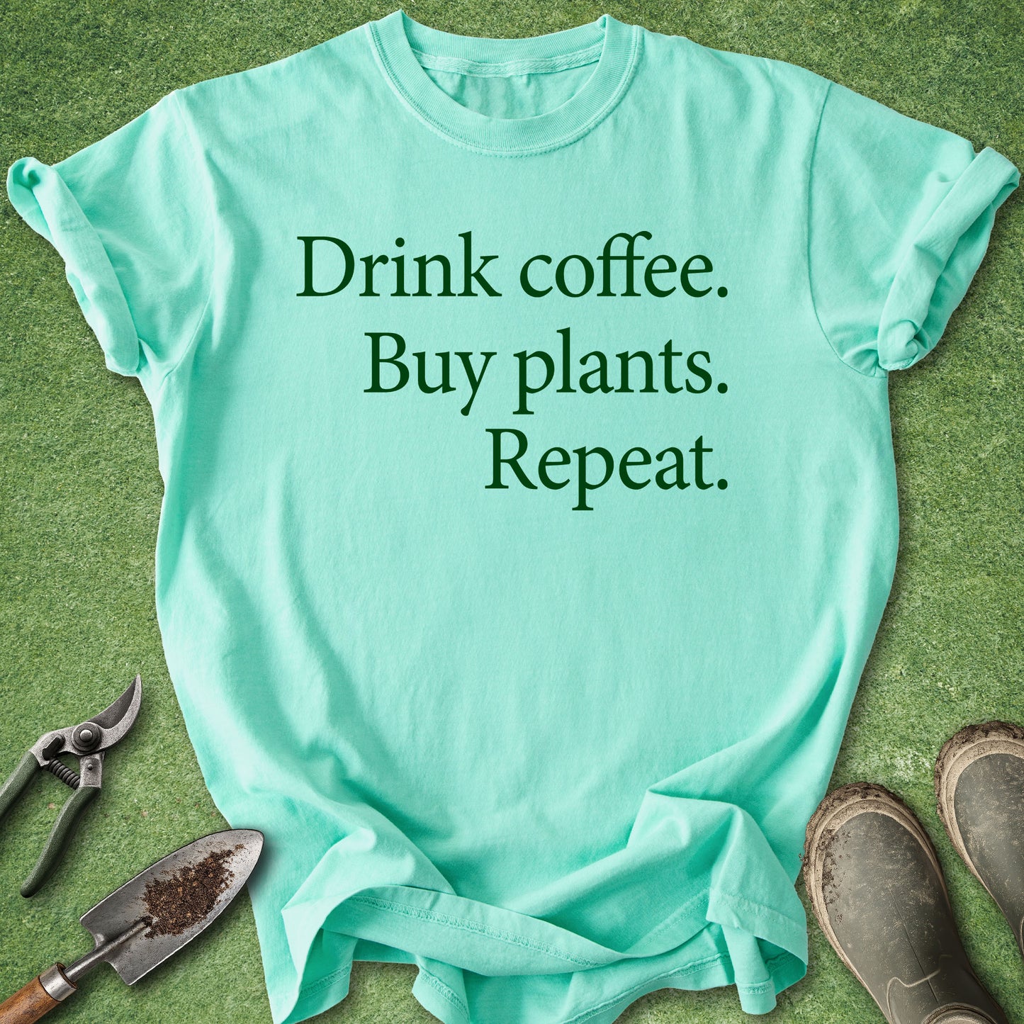 A mint green graphic t-shirt laid flat on a green grass-like surface, with gardening tools and boots around it. The shirt has the text 'Drink coffee.