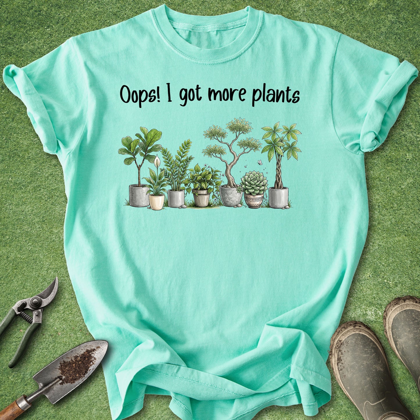 A mint green graphic t-shirt laid flat on a green grass-like surface, with gardening tools and boots around it. The shirt has black text reading 'Oops! I got more plants' above a row of illustrated potted succulents and small trees.