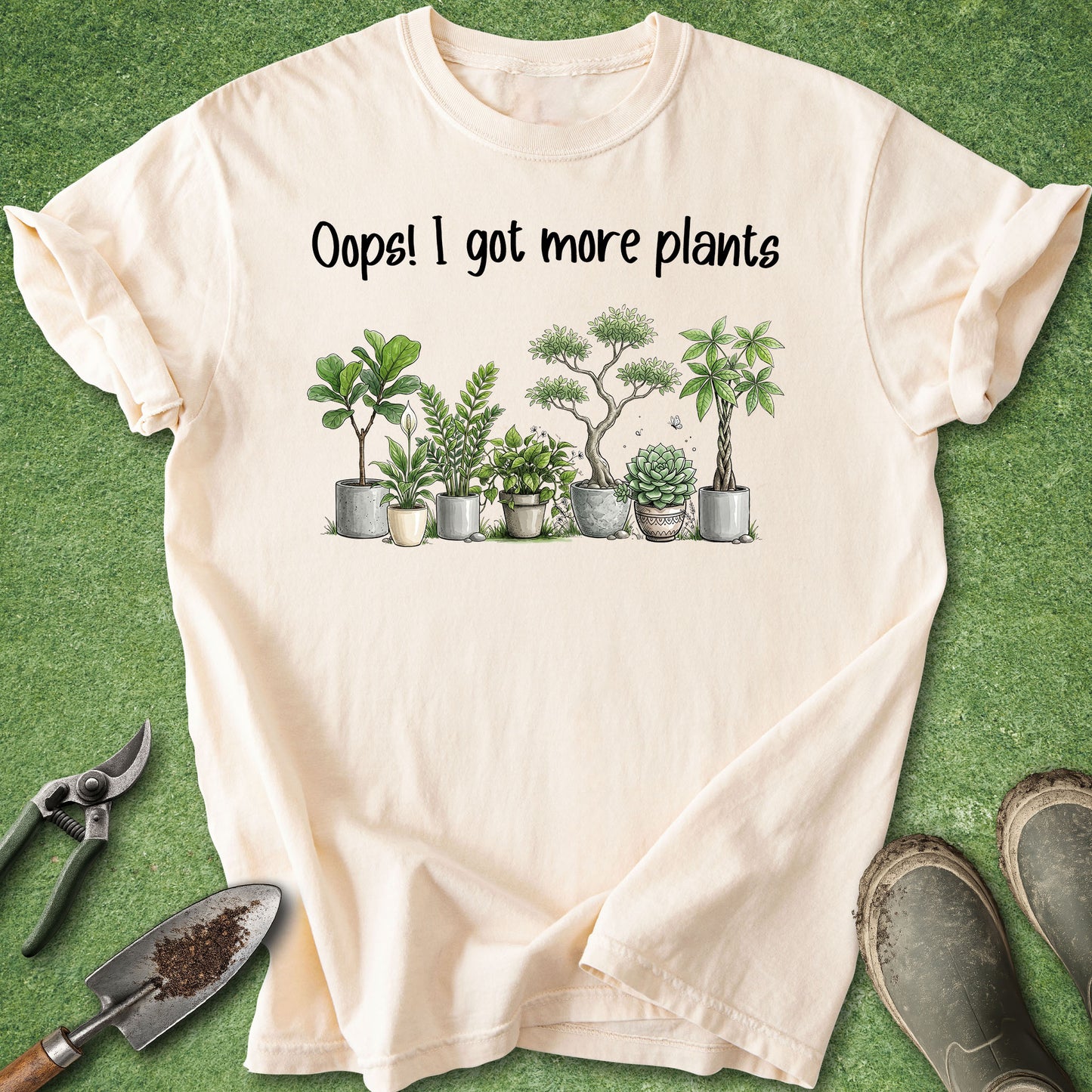 A cream-colored short-sleeve t-shirt with a graphic of various potted plants and the text 'Oops! I got more plants' printed on the chest. The shirt is laid flat on a green grass-like surface, surrounded by gardening props including pruning shears, a trowel, and gardening gloves.