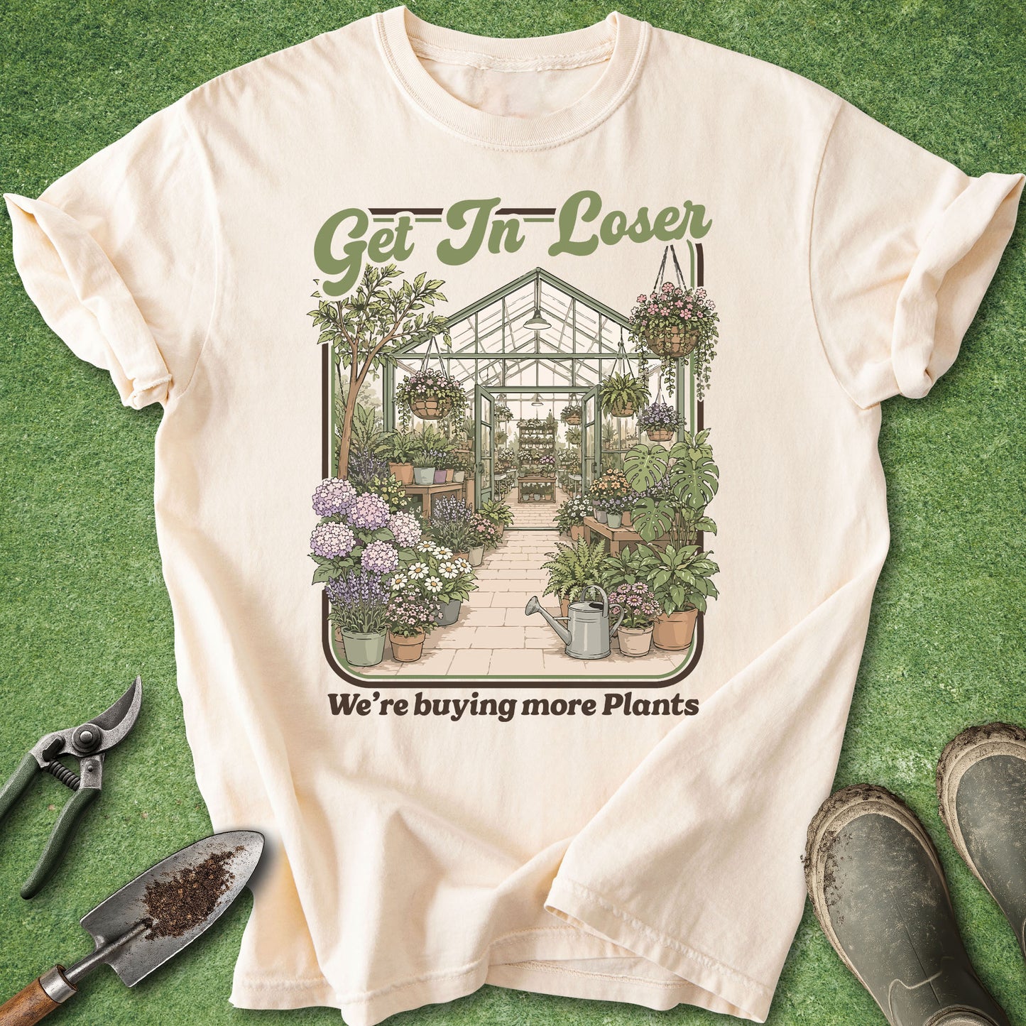 A cream-colored short-sleeve t-shirt with a green and brown graphic of a greenhouse filled with plants, laid flat on a green grass-like background with gardening tools and boots nearby.