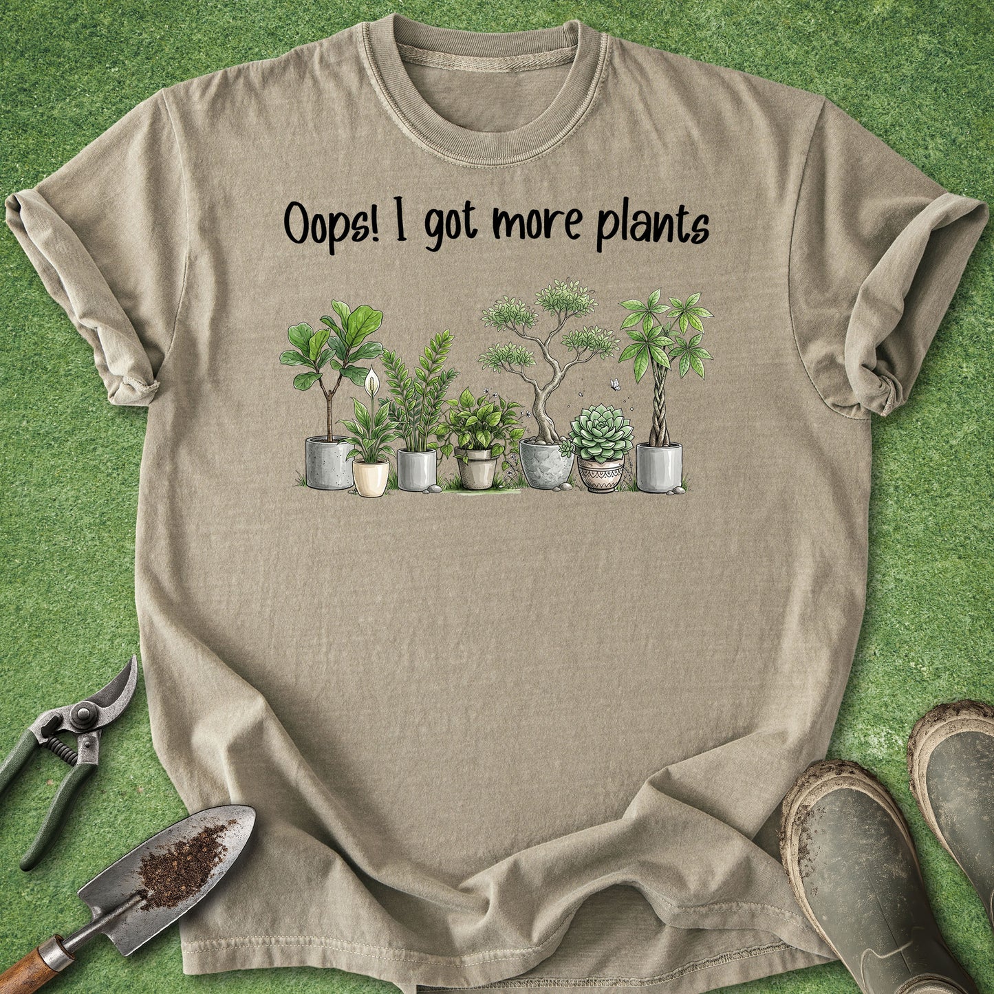 A beige short-sleeve t-shirt laid flat on a green grass-like surface, printed with the phrase 'Oops! I got more plants' above a row of illustrated potted succulents and small plants. Gardening props including pruning shears, a trowel, and boots are partially visible around the shirt.