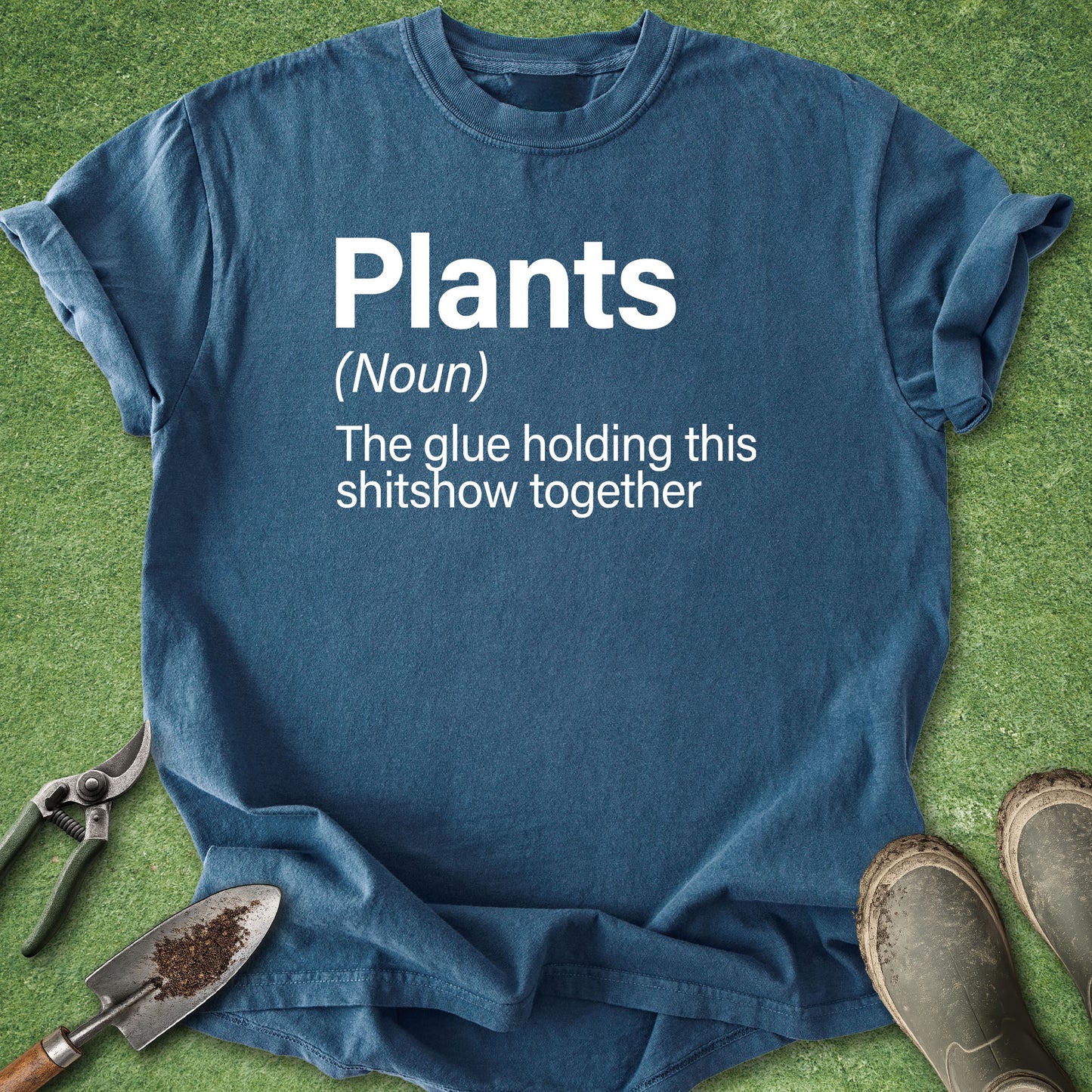 A blue graphic t-shirt laid flat on green grass with gardening tools and boots around it. The shirt has white text that reads: 'Plants (Noun) The glue holding this shitshow together'.