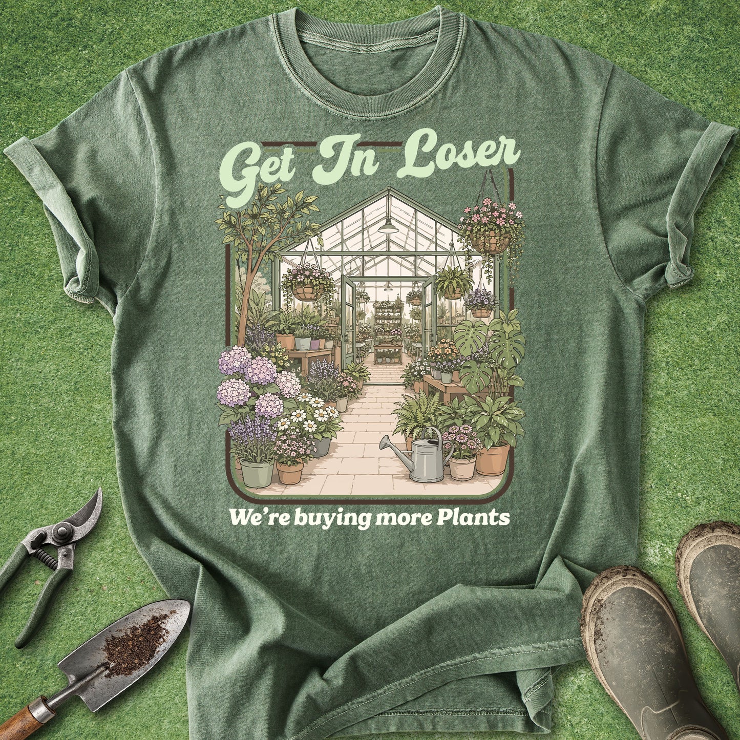 A green graphic t-shirt laid flat on a grass-like surface, featuring a illustrated greenhouse scene with plants and the text 'Get In Loser' and 'We're buying more Plants'. Gardening props including pruning shears, a trowel, and boots are placed around the shirt.