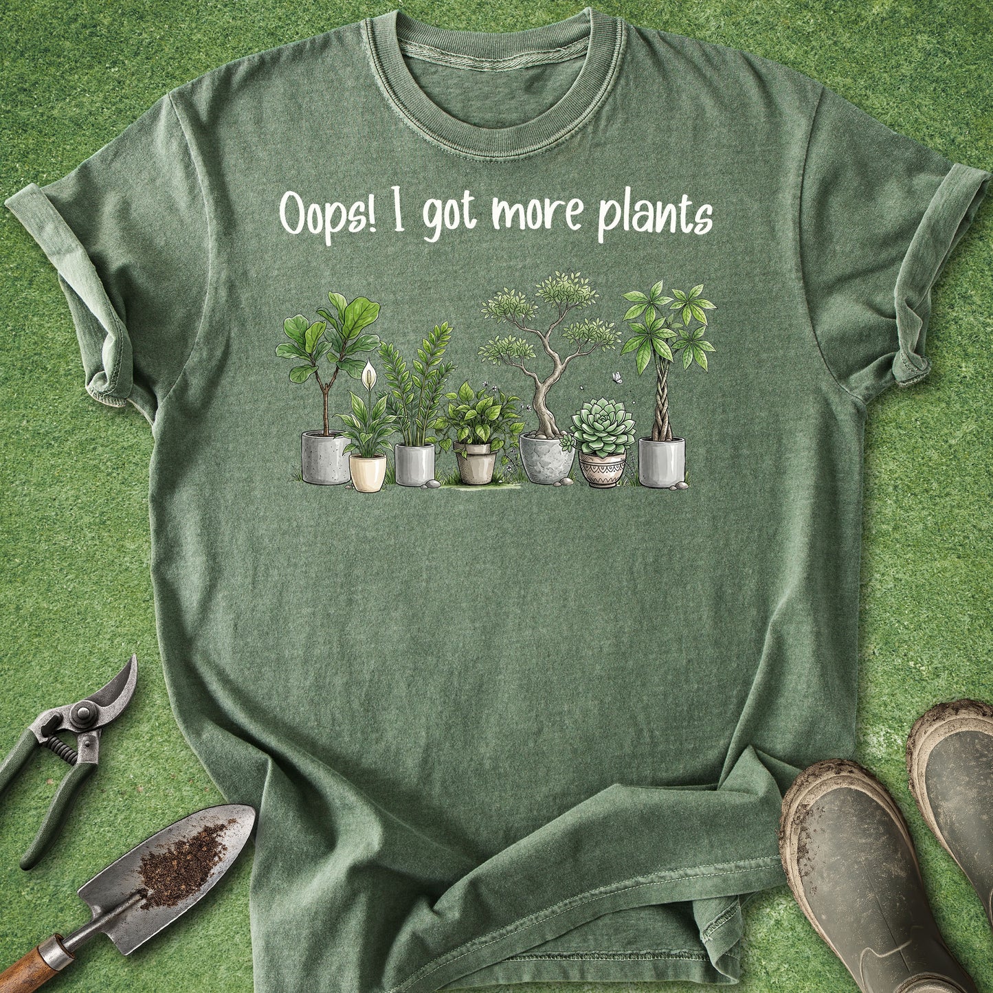 An olive green graphic t-shirt laid flat on a green grass-like surface, with gardening props including pruning shears, a trowel, and boots around it. The shirt reads 'Oops! I got more plants' above a row of illustrated potted succulents and small trees.