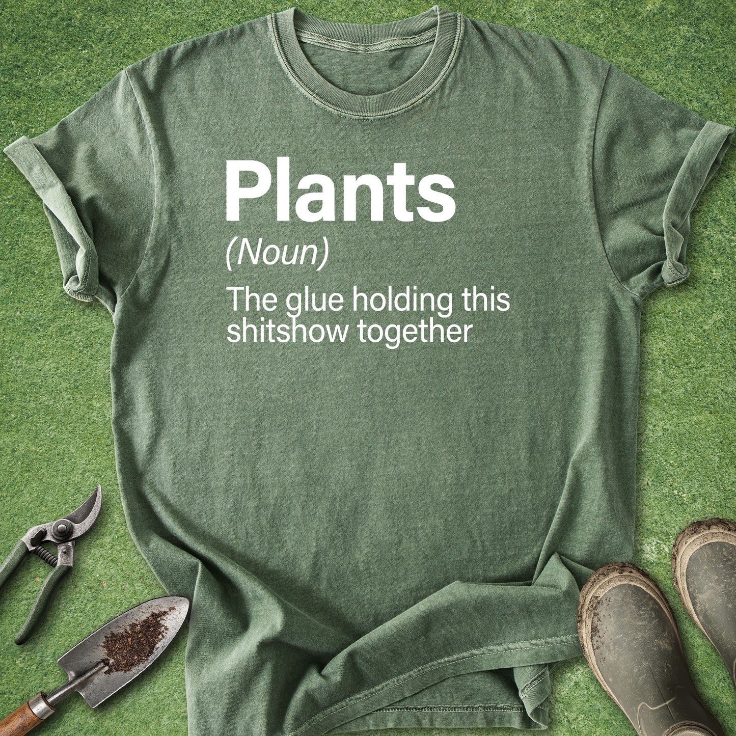 An olive green graphic t-shirt laid flat on green grass-like artificial turf, with gardening tools (shears, trowel) and boots as props. The shirt features a white printed dictionary definition of the word 'Plants' with a humorous definition.