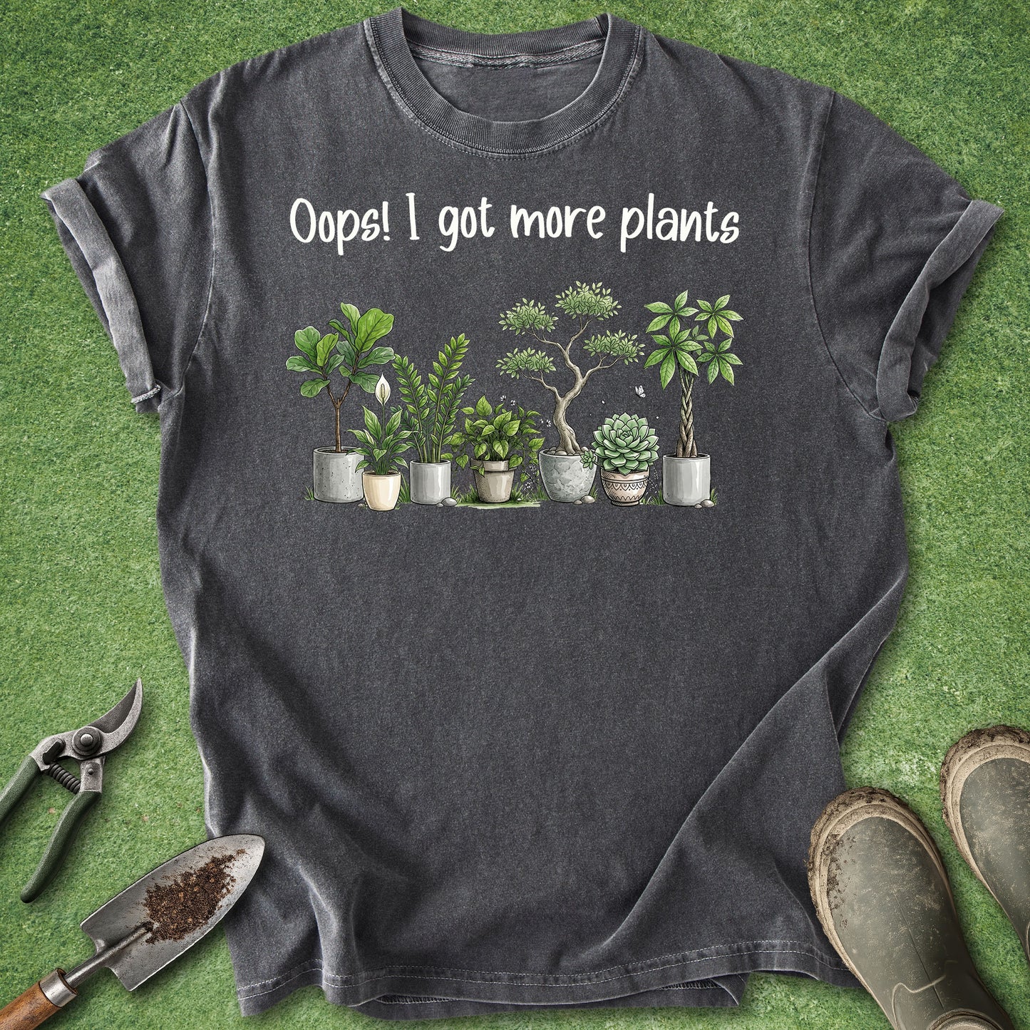 A dark gray heather t-shirt laid flat on artificial green grass, featuring the white text 'Oops! I got more plants' above a row of illustrated potted houseplants. Gardening props including pruning shears, a trowel, and boots are placed around the shirt.