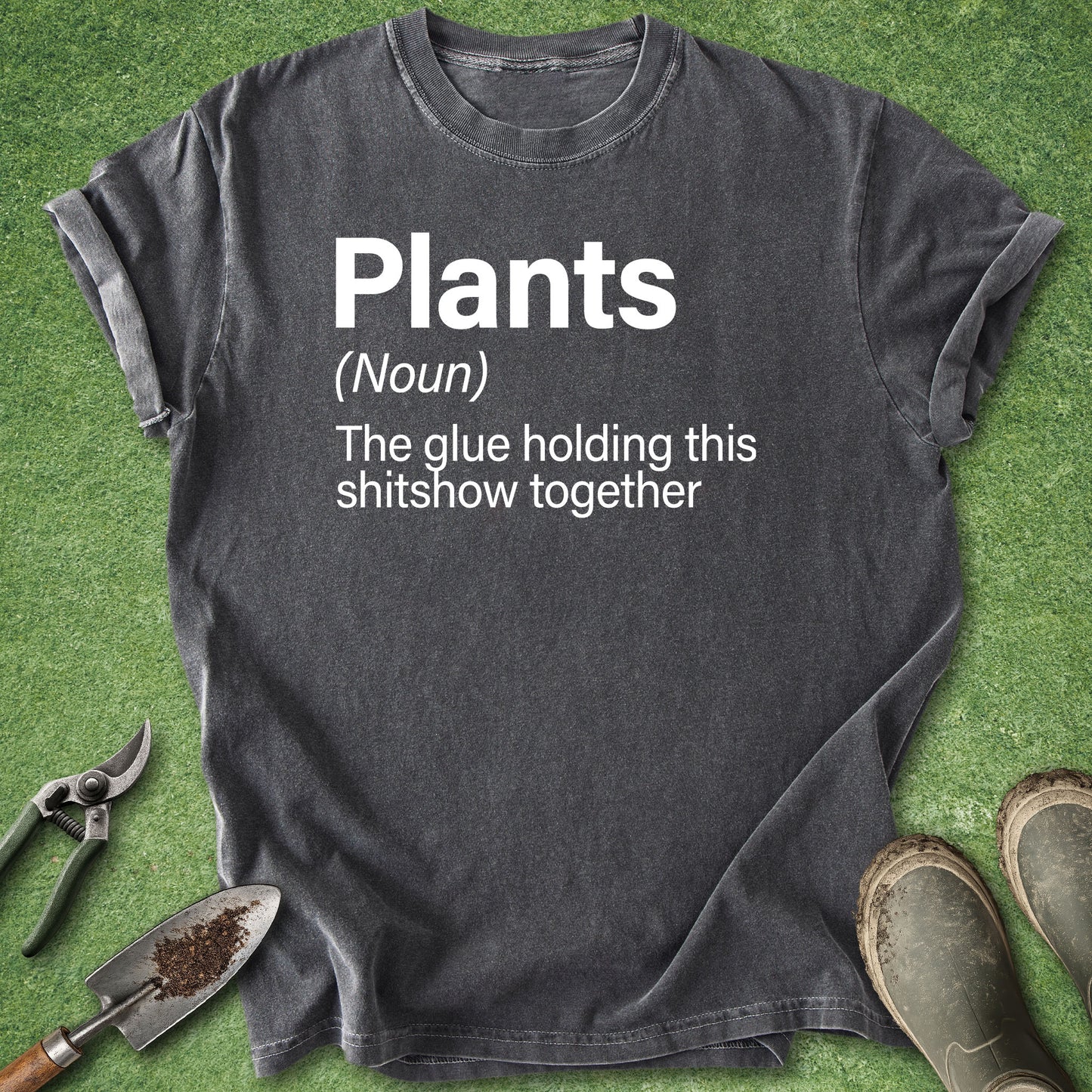 A dark charcoal gray graphic t-shirt laid flat on green artificial grass, surrounded by gardening props including pruning shears, a trowel, and boots. The shirt features a white printed dictionary definition of the word 'Plants'.