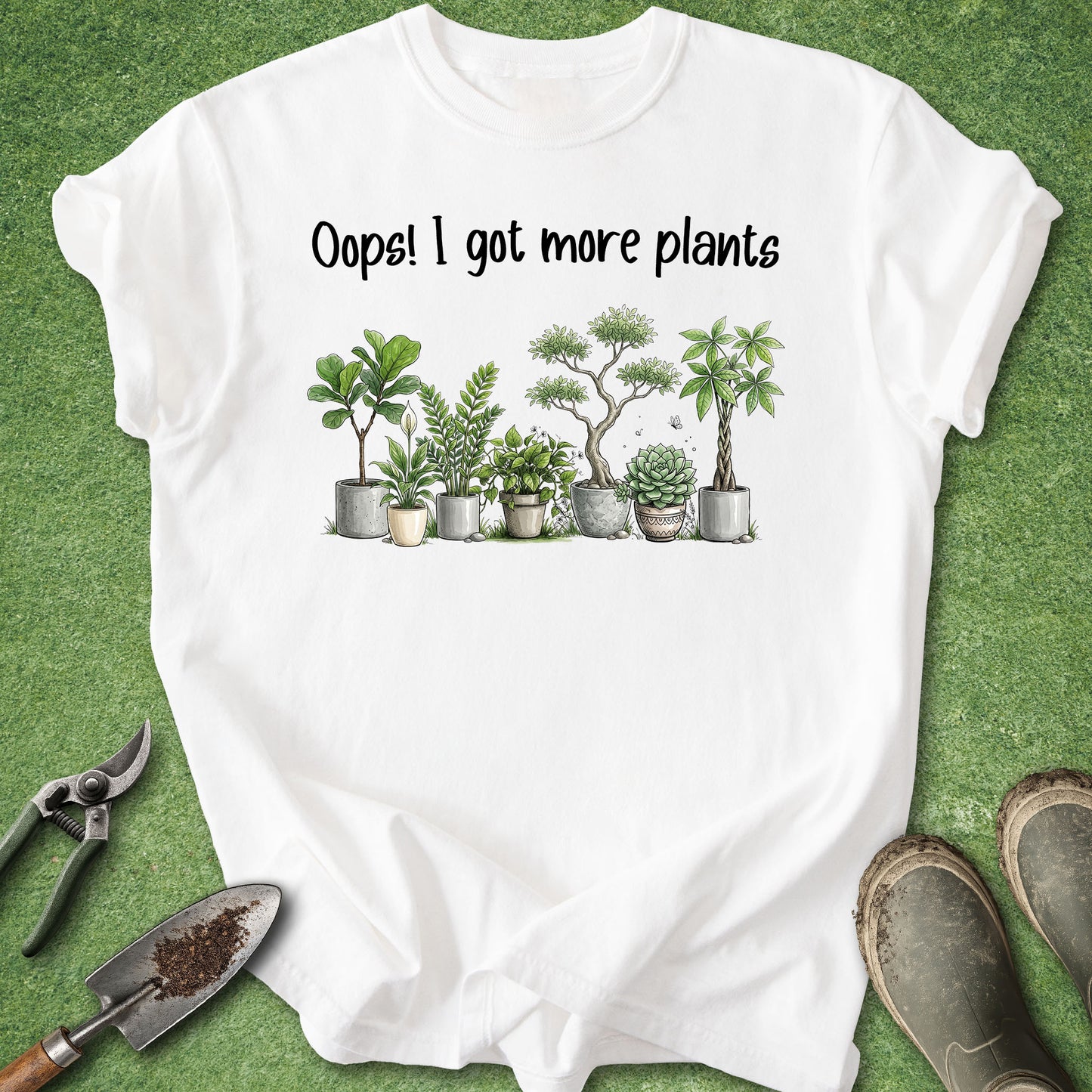 A white short-sleeve graphic T-shirt laid flat on green grass-like flooring, with the printed phrase 'Oops! I got more plants' above a row of illustrated potted succulents and small trees. Gardening props, including pruning shears, a trowel, and boots, are visible around the shirt.