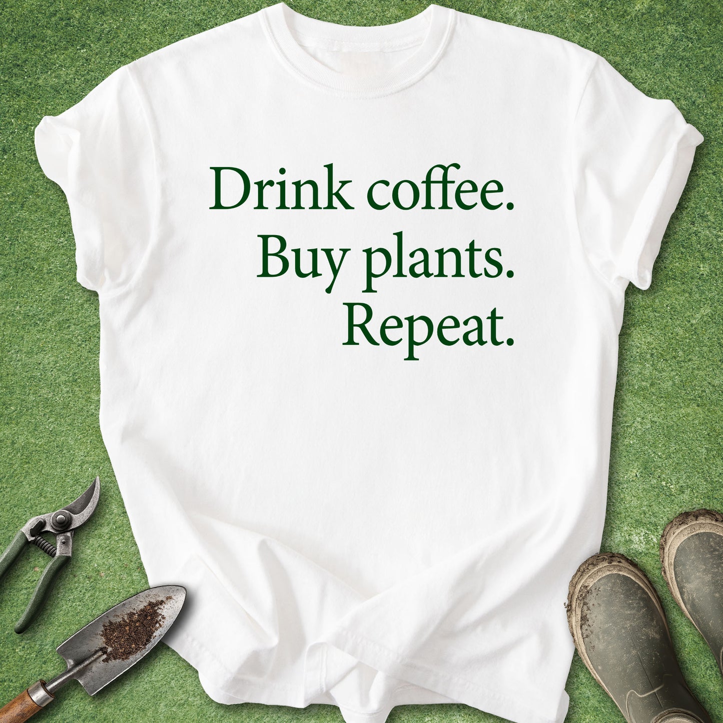 A white short-sleeve T-shirt laid flat on green grass, printed with the phrase 'Drink coffee. Buy plants.