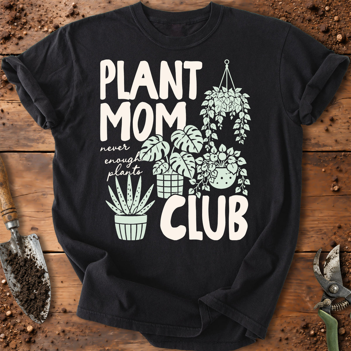 A black short-sleeve graphic T-shirt laid flat on a rustic wooden surface with gardening props. The shirt features large white text reading 'PLANT MOM CLUB' with smaller script text 'never enough plants' and illustrated plants including hanging succulents, a monstera, a potted succulent, and an aloe vera.