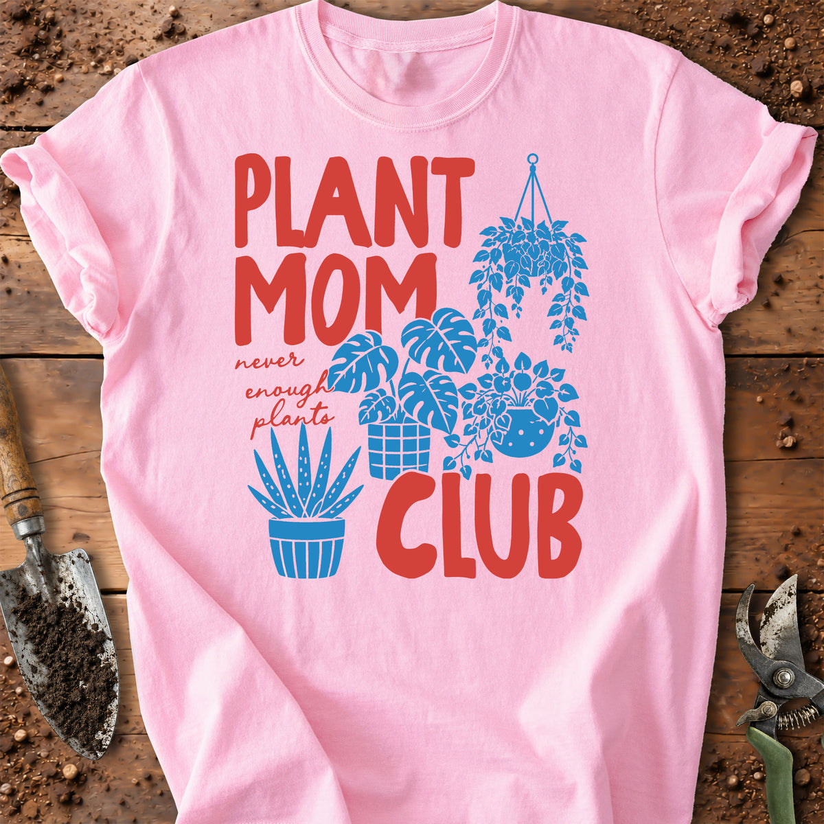 A pink short-sleeve graphic T-shirt laid flat on a rustic wooden surface with gardening props. The shirt features the phrase "PLANT MOM CLUB" in large red letters, with smaller red text reading "never enough plants" and blue plant illustrations including a hanging plant, potted plants, and an aloe vera.