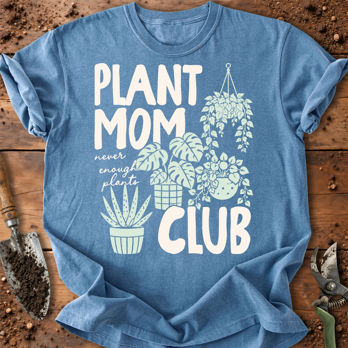 A blue graphic T-shirt laid flat on a rustic wooden surface with gardening props. The shirt reads “PLANT MOM never enough plants CLUB” in large white text, with illustrated potted and hanging plants in light blue.