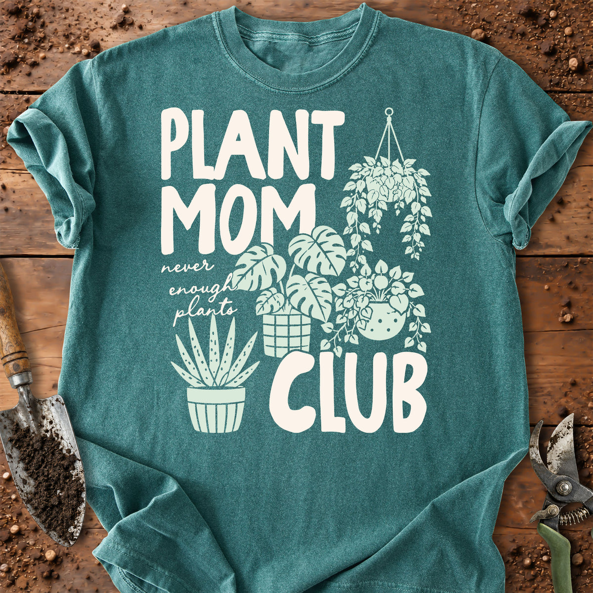 A teal short-sleeve graphic T-shirt laid flat on a rustic wooden surface with gardening props. The shirt has a large white printed design reading “PLANT MOM CLUB” with illustrated potted and hanging plants and the phrase “never enough plants.