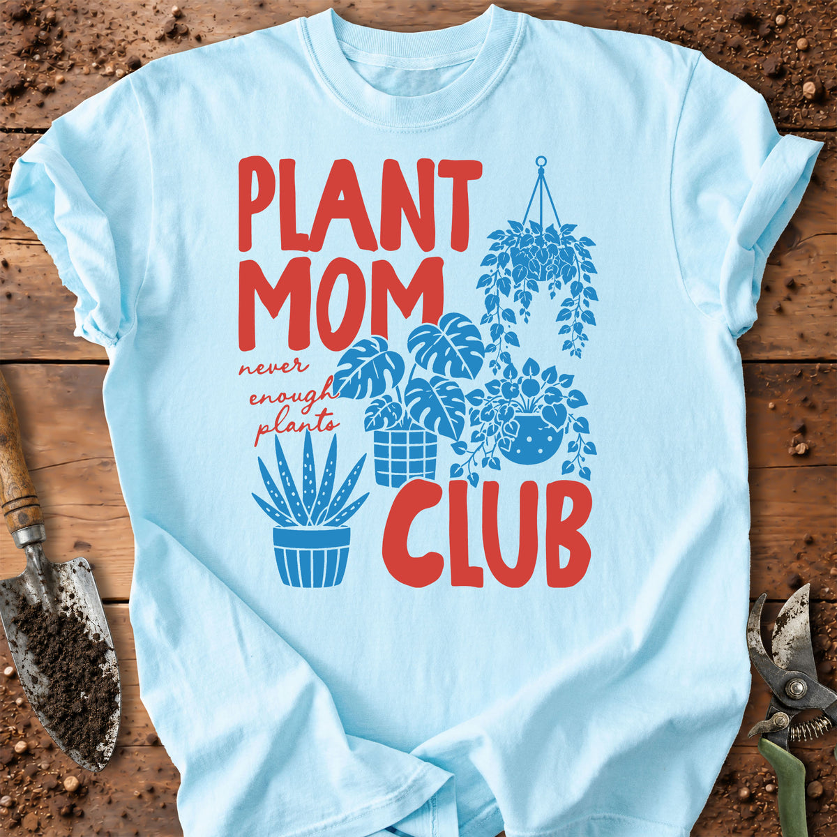 A light blue graphic t-shirt laid flat on a rustic wooden surface with gardening props. The shirt features a red and blue graphic that reads 'PLANT MOM CLUB' with illustrations of potted and hanging plants.
