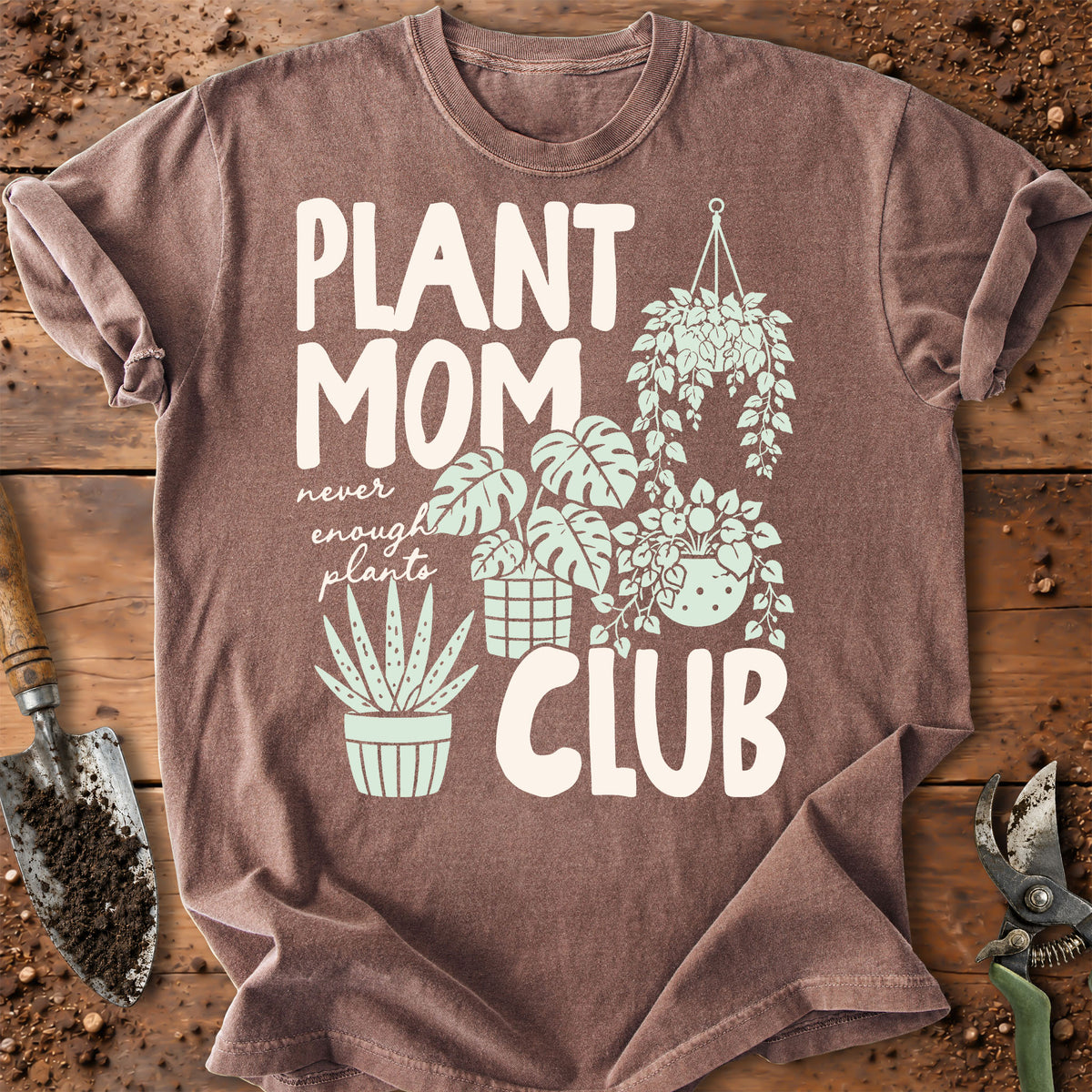 A brown short-sleeve graphic T-shirt laid flat on a rustic wooden surface with soil and gardening tools. The shirt reads “PLANT MOM CLUB” in large white letters, with smaller white script text and illustrated green plants and pots.