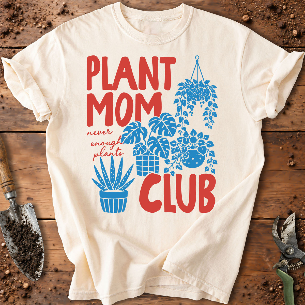 An off-white graphic t-shirt laid flat on a rustic wooden surface with gardening tools. The shirt features a colorful print with the text 'PLANT MOM CLUB' in red and blue, accompanied by illustrations of potted plants and a hanging plant.