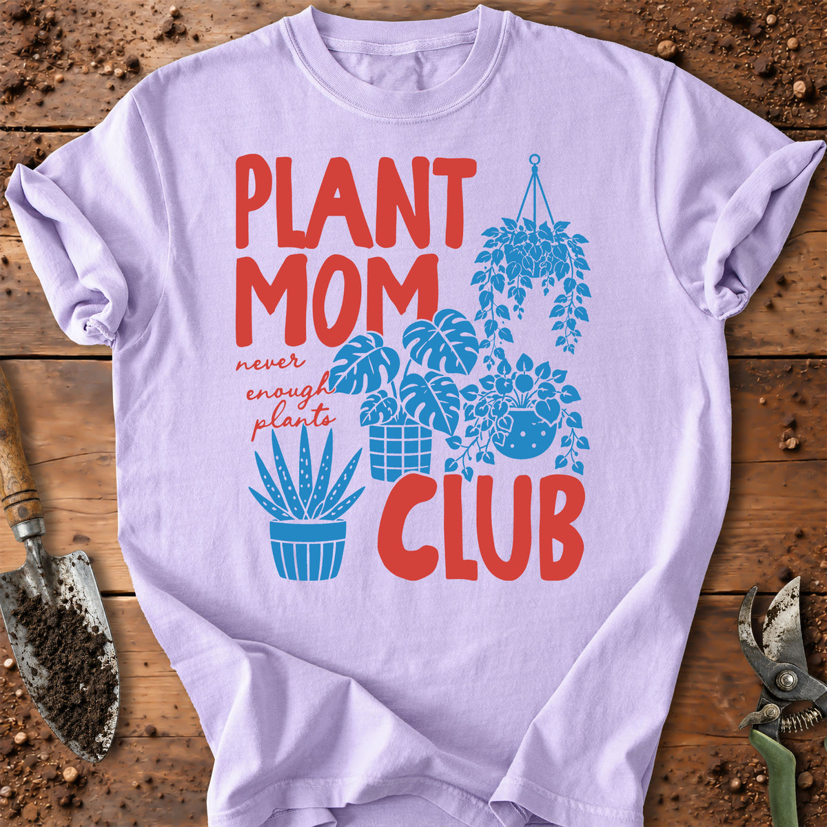 A light lavender crewneck t-shirt with a 'PLANT MOM NEVER ENOUGH PLANTS CLUB' graphic in red and blue, laid flat on a rustic wooden background with gardening tools.