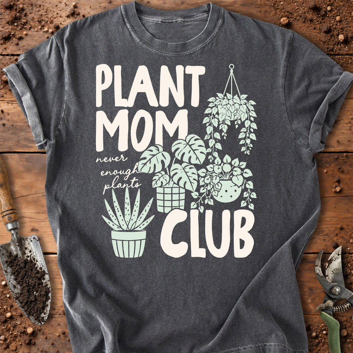 A dark charcoal gray graphic t-shirt laid flat on a rustic wooden surface with gardening props. The shirt features a white and mint green graphic that reads 'PLANT MOM never enough plants CLUB' alongside illustrations of potted and hanging plants.