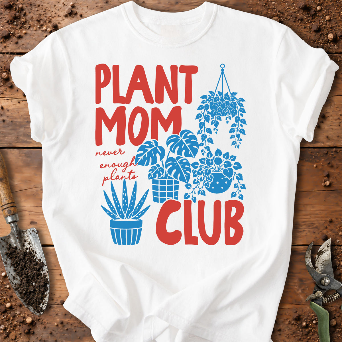 A white short-sleeve T-shirt laid flat on a rustic wooden surface with gardening props. The shirt features a colorful graphic that reads 'PLANT MOM CLUB' in red and blue, with illustrated potted and hanging plants and small cursive text reading 'never enough plants.