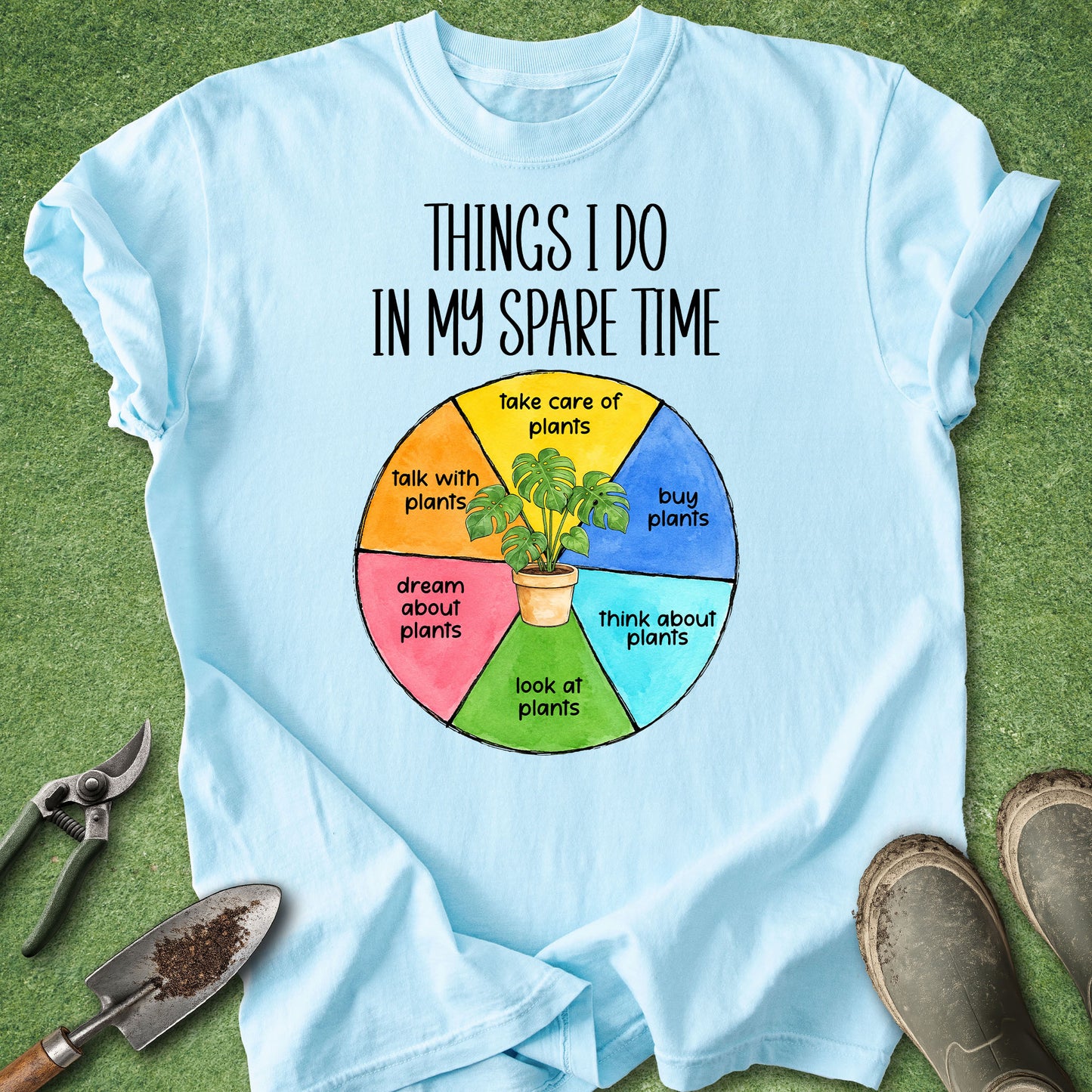 A light blue graphic t-shirt laid flat on green grass with gardening props like pruning shears, a trowel, and boots around it. The shirt features a colorful wheel graphic and the text 'THINGS I DO IN MY SPARE TIME' with plant-related activities listed in the wheel segments.