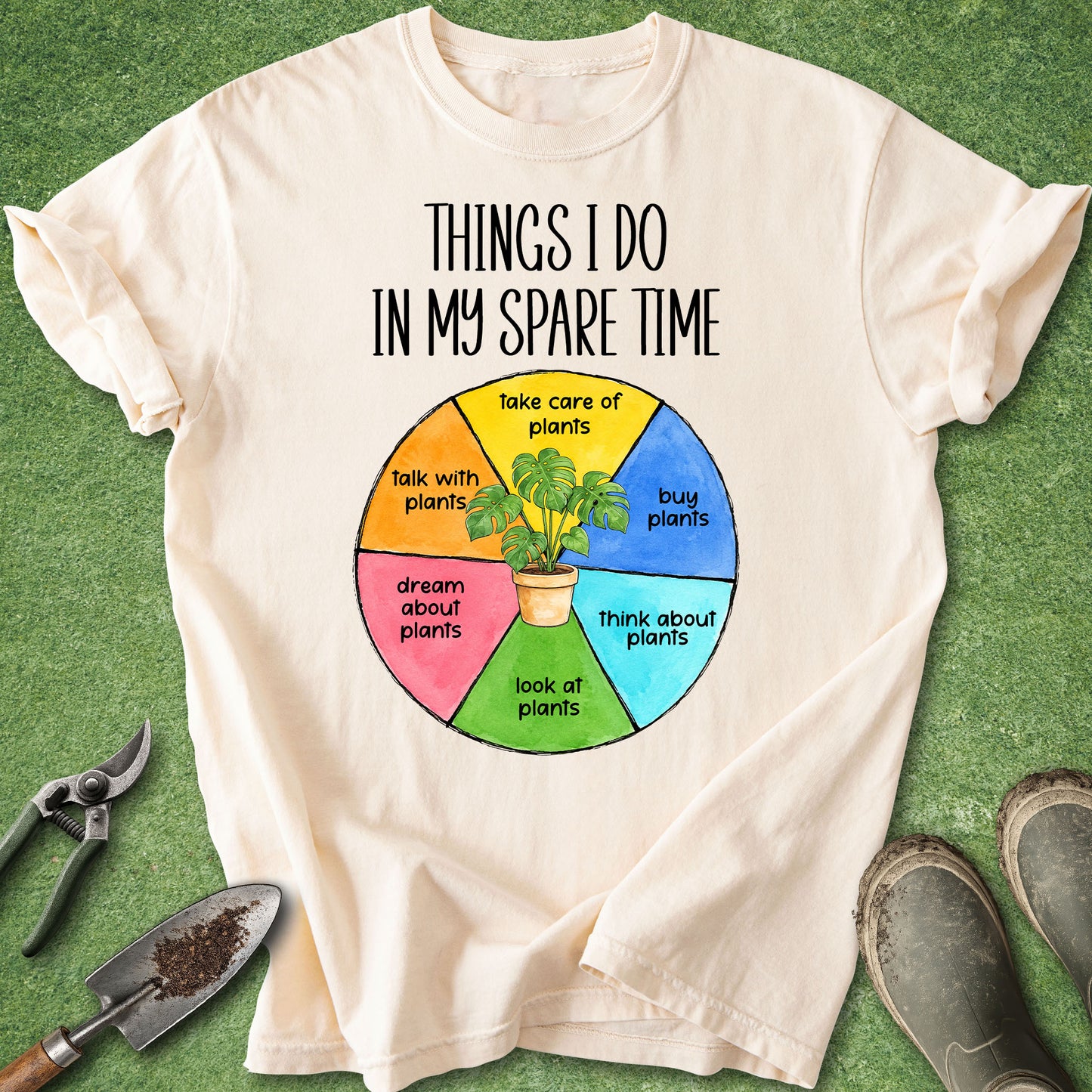 A cream-colored graphic t-shirt laid flat on green grass with gardening tools and shoes around it. The shirt features a colorful circular diagram with the text 'THINGS I DO IN MY SPARE TIME' and sections like 'take care of plants', 'talk with plants', 'buy plants', 'dream about plants', 'look at plants', and 'think about plants'.