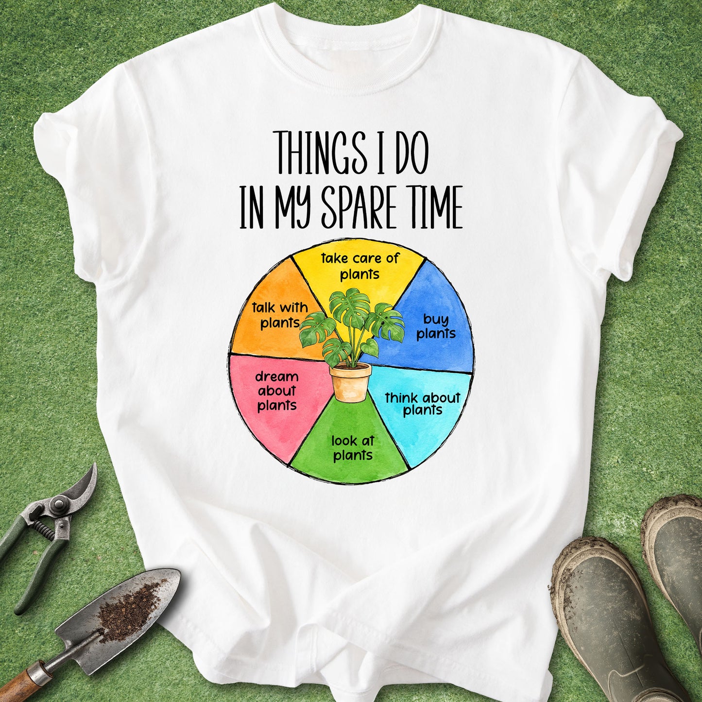 A white graphic T-shirt laid flat on green grass with gardening props around it. The shirt has black text reading 'THINGS I DO IN MY SPARE TIME' above a colorful circular diagram with plant-related phrases and a small potted plant illustration.