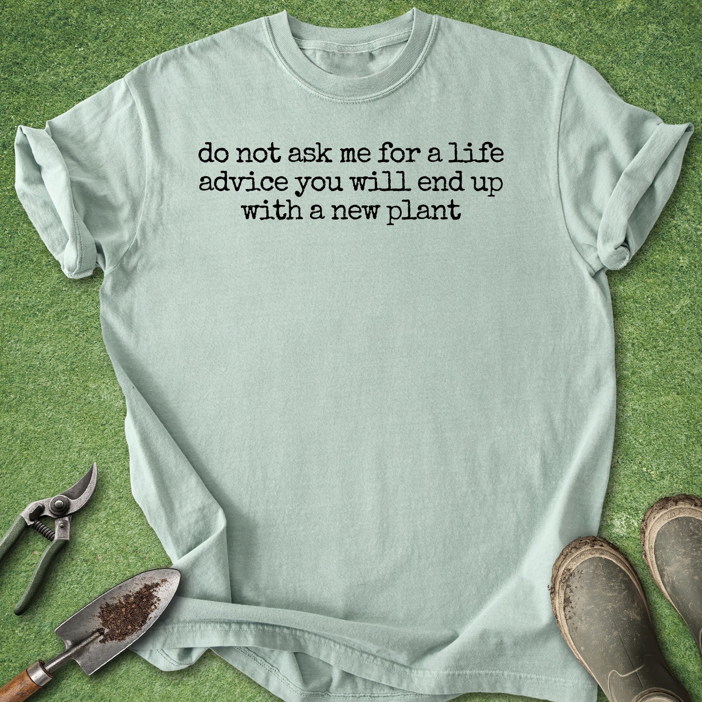 A light sage green t-shirt with black printed text reading 'do not ask me for a life advice you will end up with a new plant' laid flat on a green grass-like surface, styled with gardening tools and muddy boots.